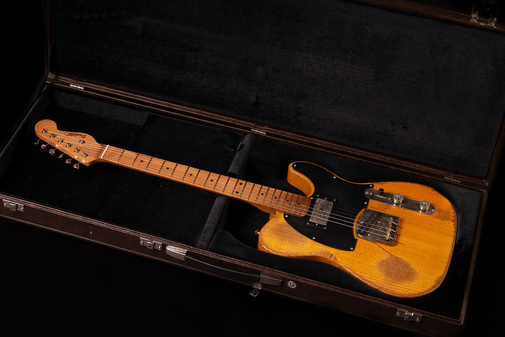 An electric guitar with an aged chestnut wood body in butterscotch coloring in a brown leather hard case