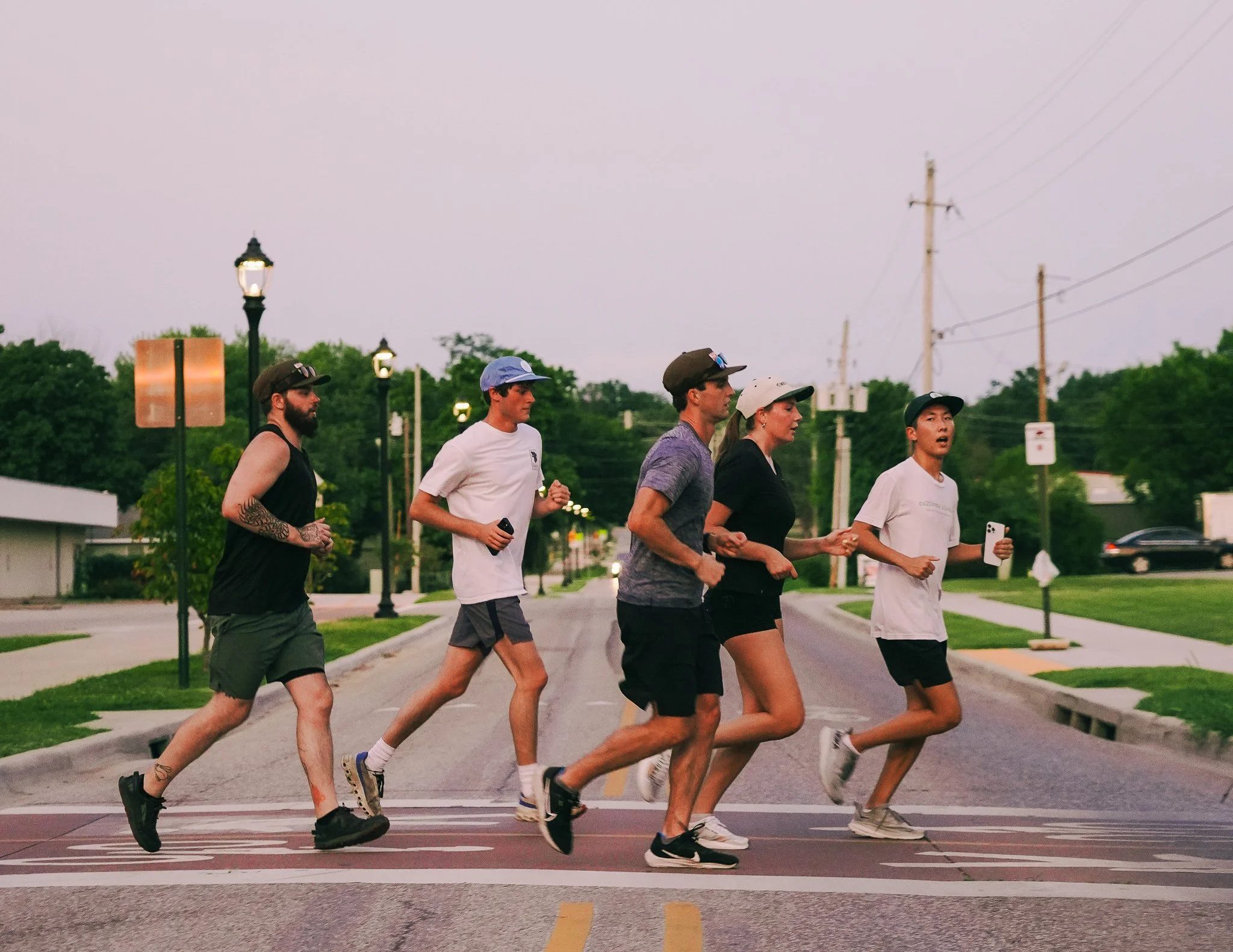 Five people running across a crosswalk on a street in a suburban area during dusk.