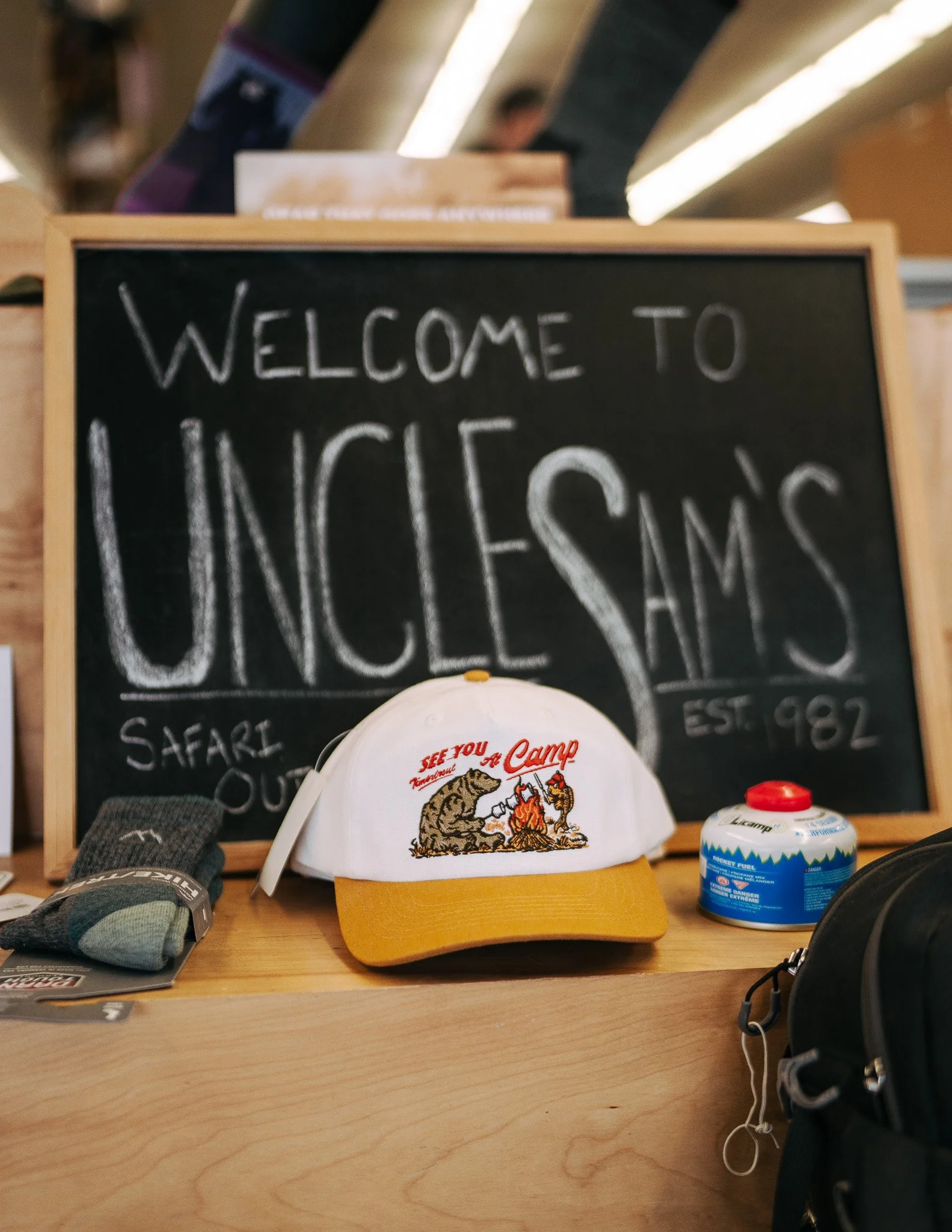 Wooden counter featuring a 'Welcome to Uncle Sam's' chalkboard, a white TimberTrout Campfire Snapback hat, and camping accessories.