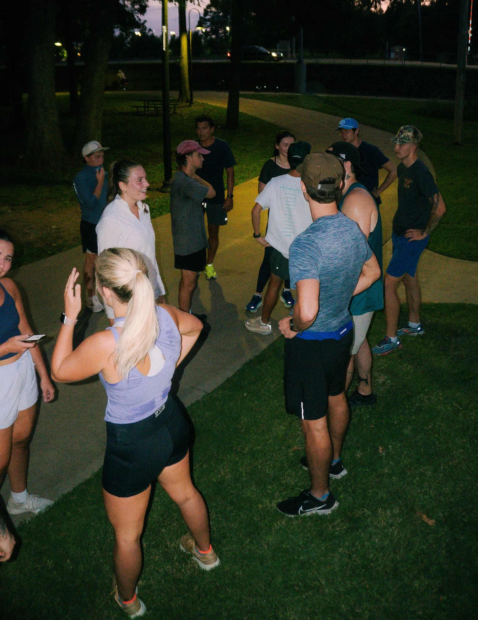 Members of the TimberTrout Run Club gathered on a park sidewalk at dusk wearing athletic gear and engaging in conversation.