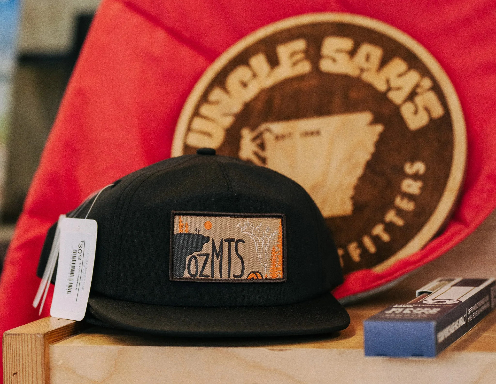 Close-up of a black TimberTrout Ozark Snapback with a rectangular landscape patch on a wooden surface with a red cloth.