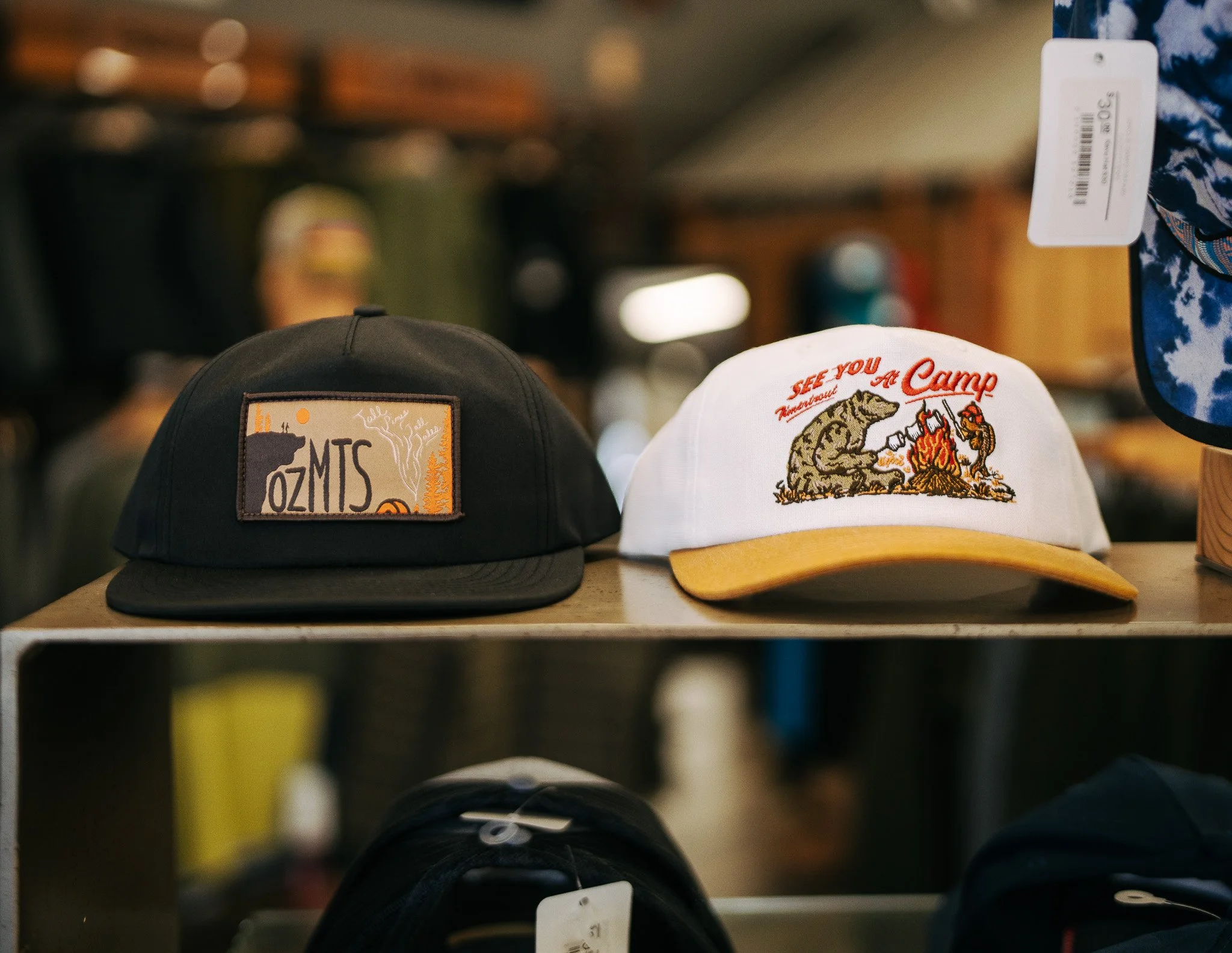 Two TimberTrout hats on display: a black Ozark Snapback with a rectangular patch and a white Campfire Snapback with a yellow brim.