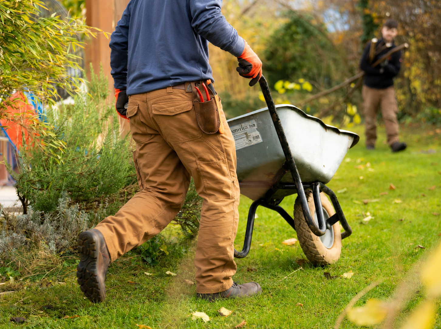 Person pushing a wheelbarrow near plants in a garden, with a child in the background holding a rake.