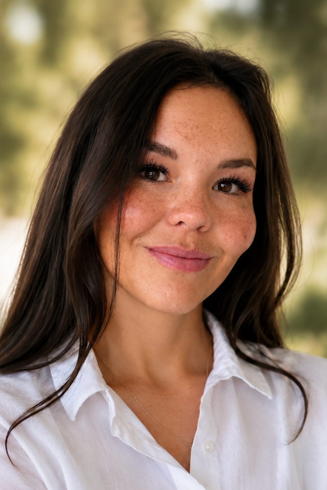 A woman with long dark hair, freckles, and a white shirt, smiling outdoors with a blurred background of trees and greenery.