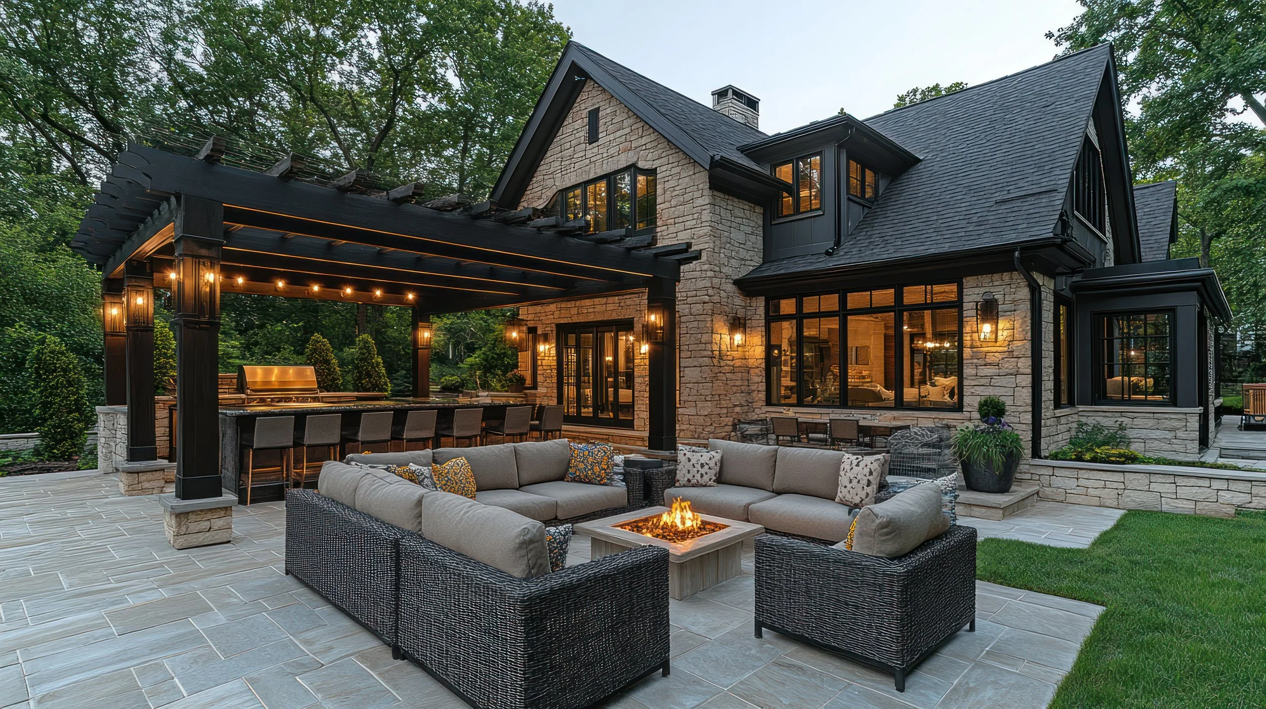 A modern house with stone exterior walls and dark roofing, featuring large windows and a spacious outdoor patio with furniture, a fire pit, and a shaded area with string lights, surrounded by lush green trees.
