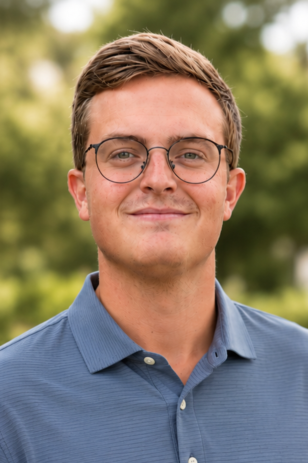 A young man with light skin, brown hair, glasses, and a slight smile, standing outdoors in front of blurred green trees, wearing a blue collared shirt.