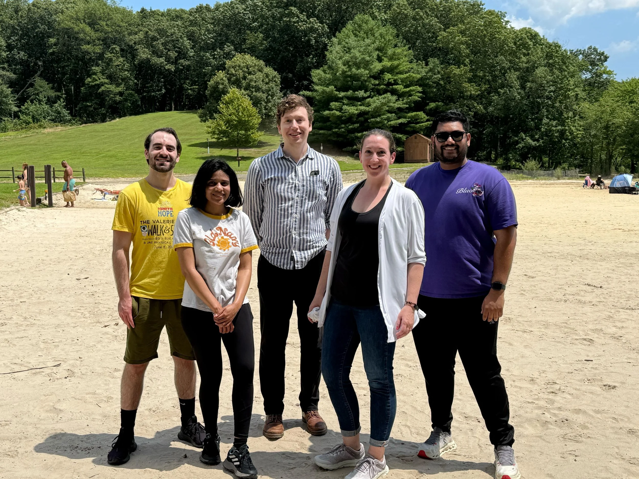 Group of five young adults standing together on a sandy beach, with a backdrop of green trees and a small wooden shed. They are smiling and dressed casually.