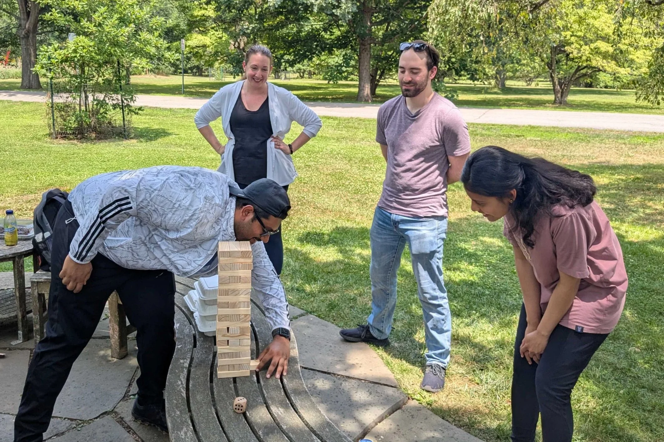 Four people gathered around a giant Jenga game on a park bench, with one person making a move, while the others watch and laugh in a sunny park with green trees in the background.