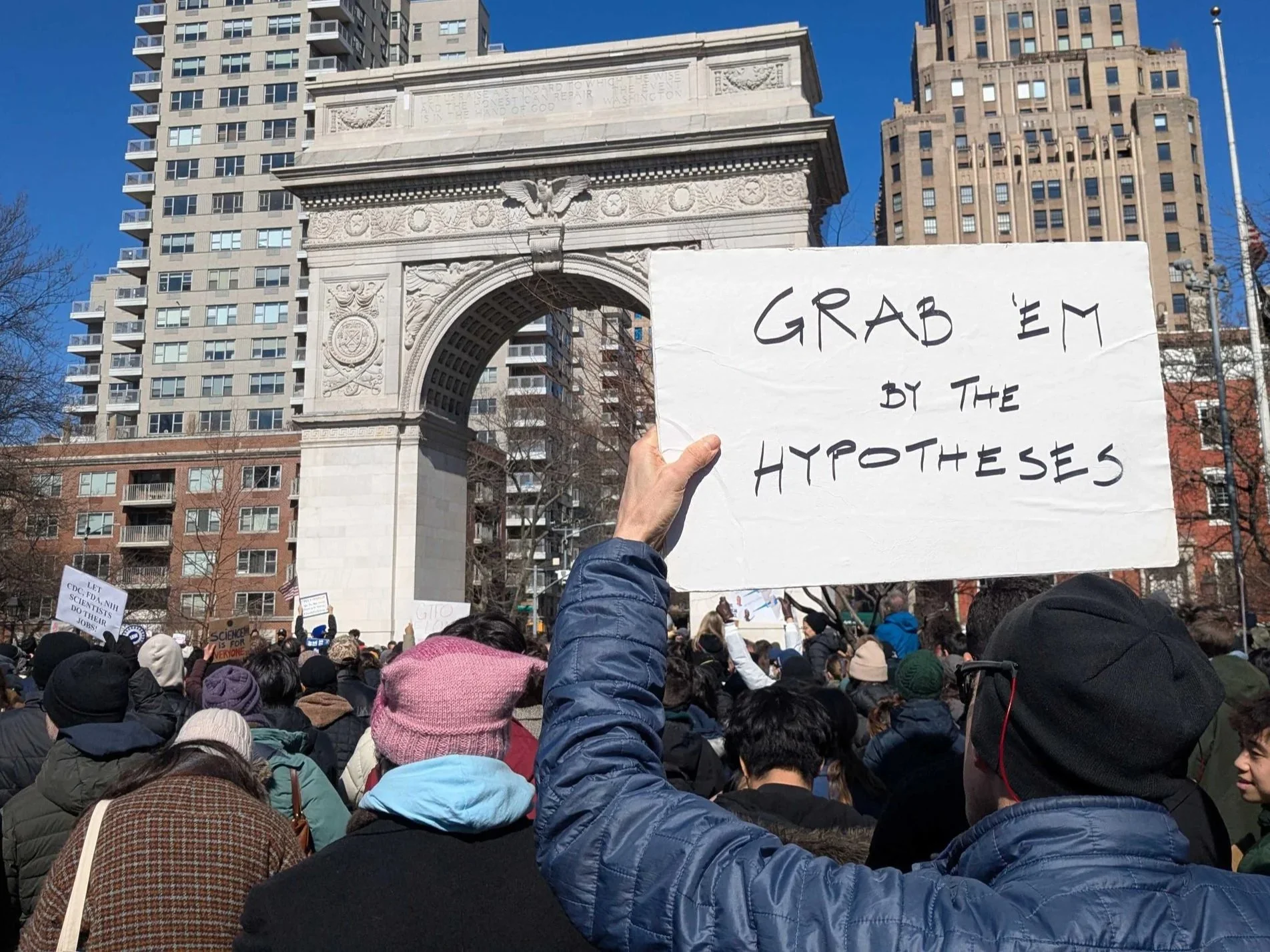 A crowd of people gathered at a protest or rally outdoors on a sunny day, with skyscrapers and a historical arch in the background. One person holds a white sign that reads 'GAB EM BY THE HYPOTHESES'.