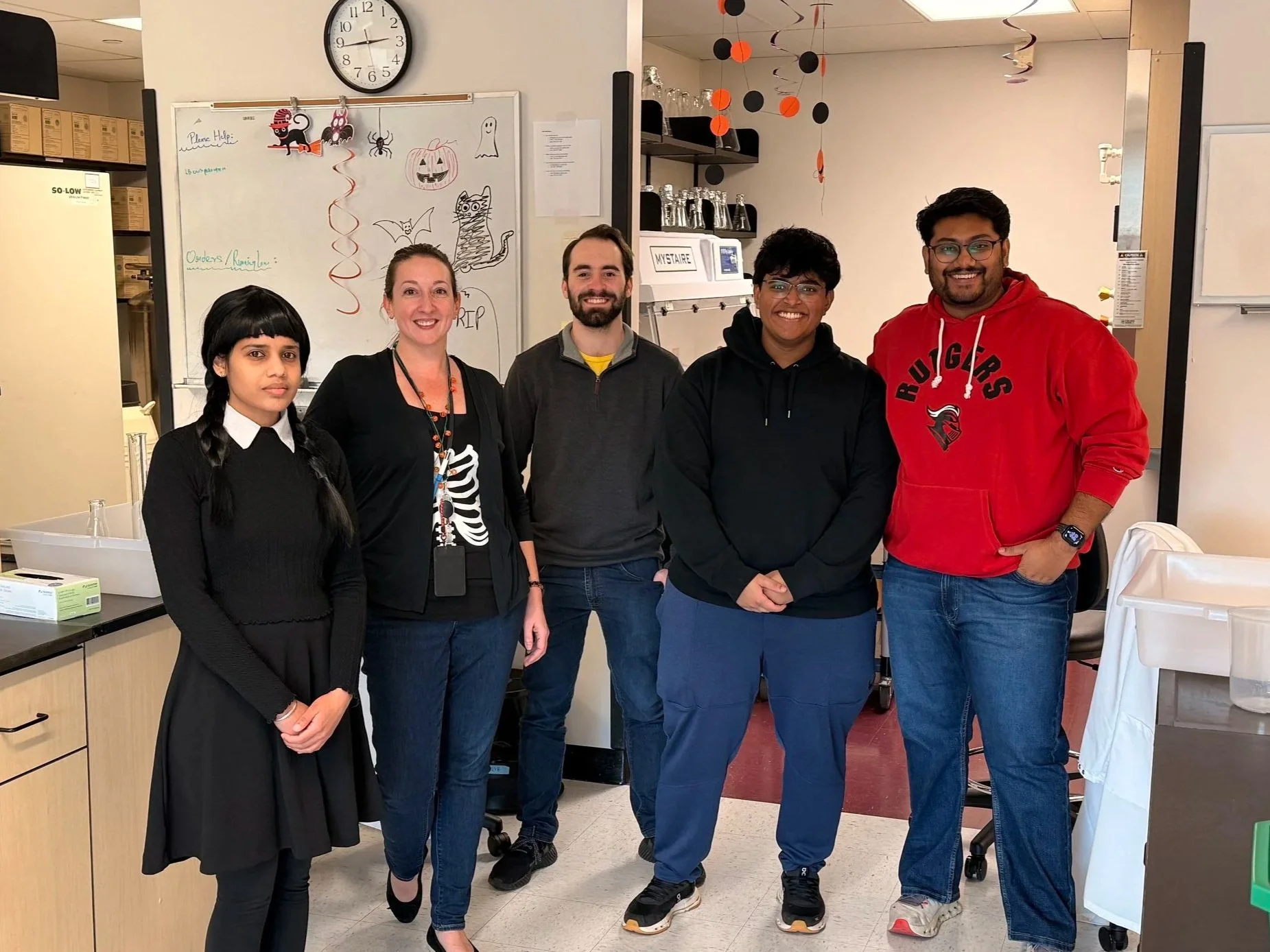 A group of five diverse people standing together in a room decorated with Halloween-themed decorations such as paper lanterns and drawings of a pumpkin, bat, and ghost on a whiteboard. They are smiling and facing the camera.