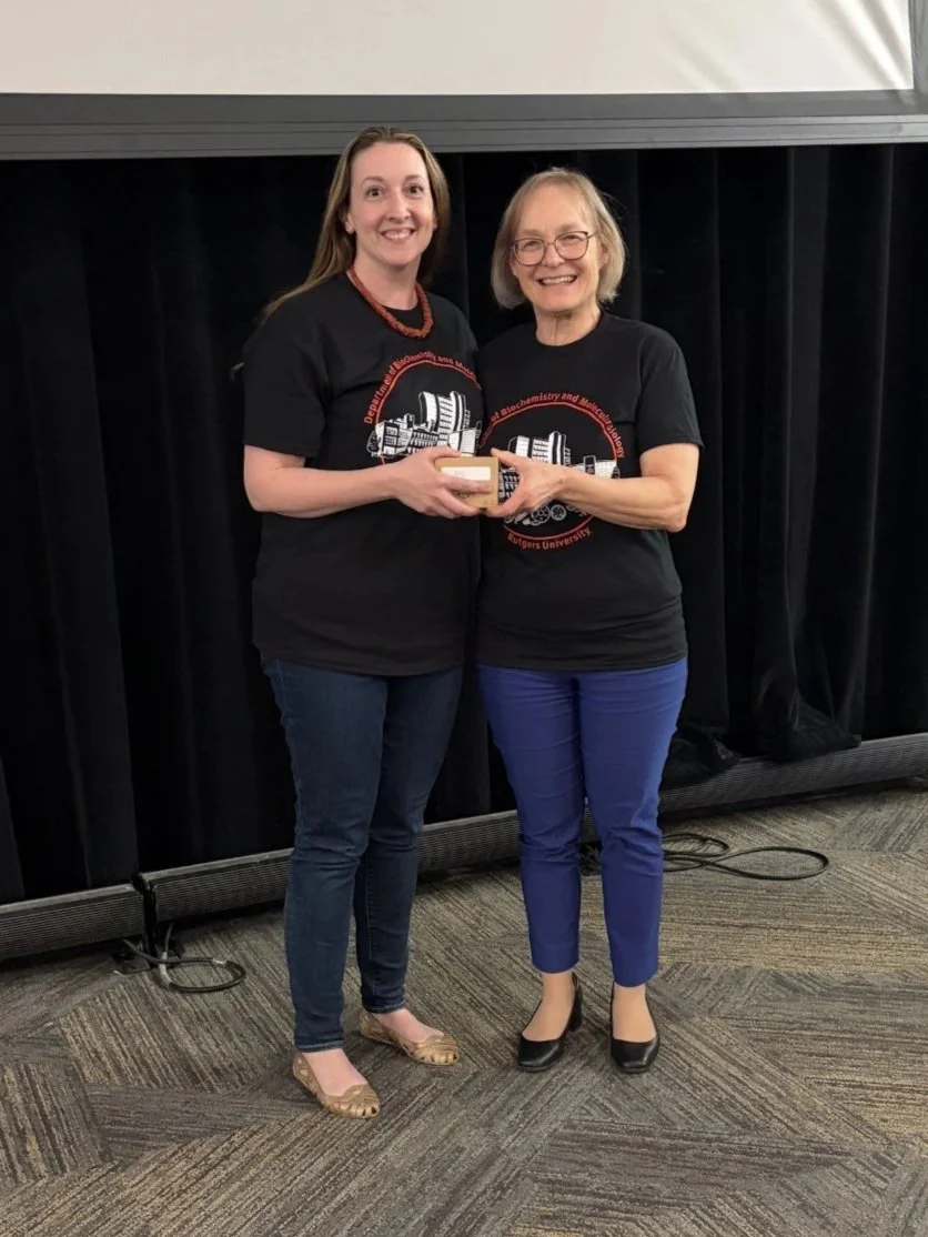 Two women standing side by side, smiling, with the woman on the right giving a small award or plaque to the woman on the left. They are both wearing black T-shirts with a design related to Rutgers University, featuring a DNA strand and a building, and are in front of black curtains and a large screen in what appears to be an event or presentation setting.