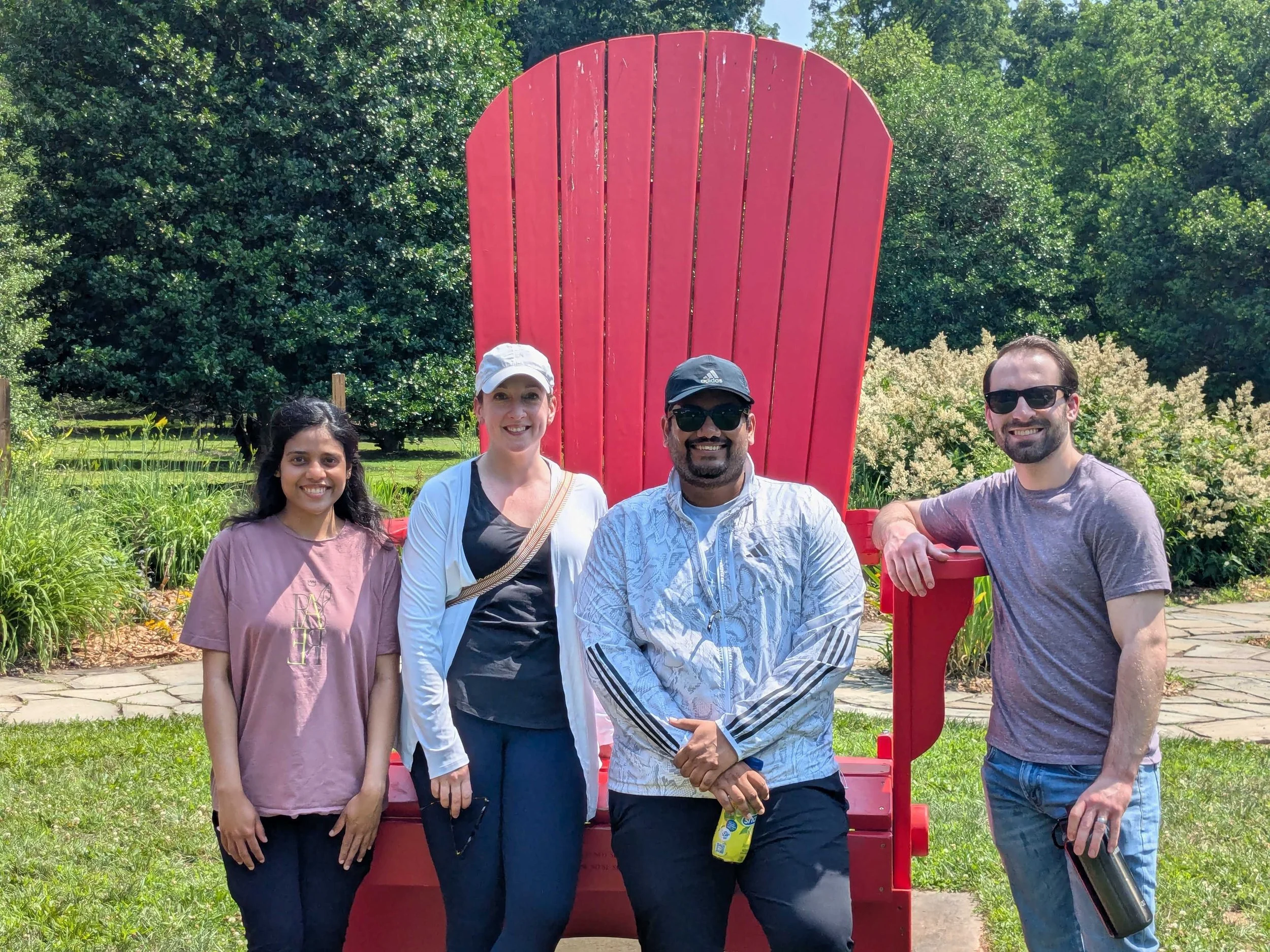 Five diverse people standing outdoors near a large red Adirondack chair with a background of green trees and bushes.