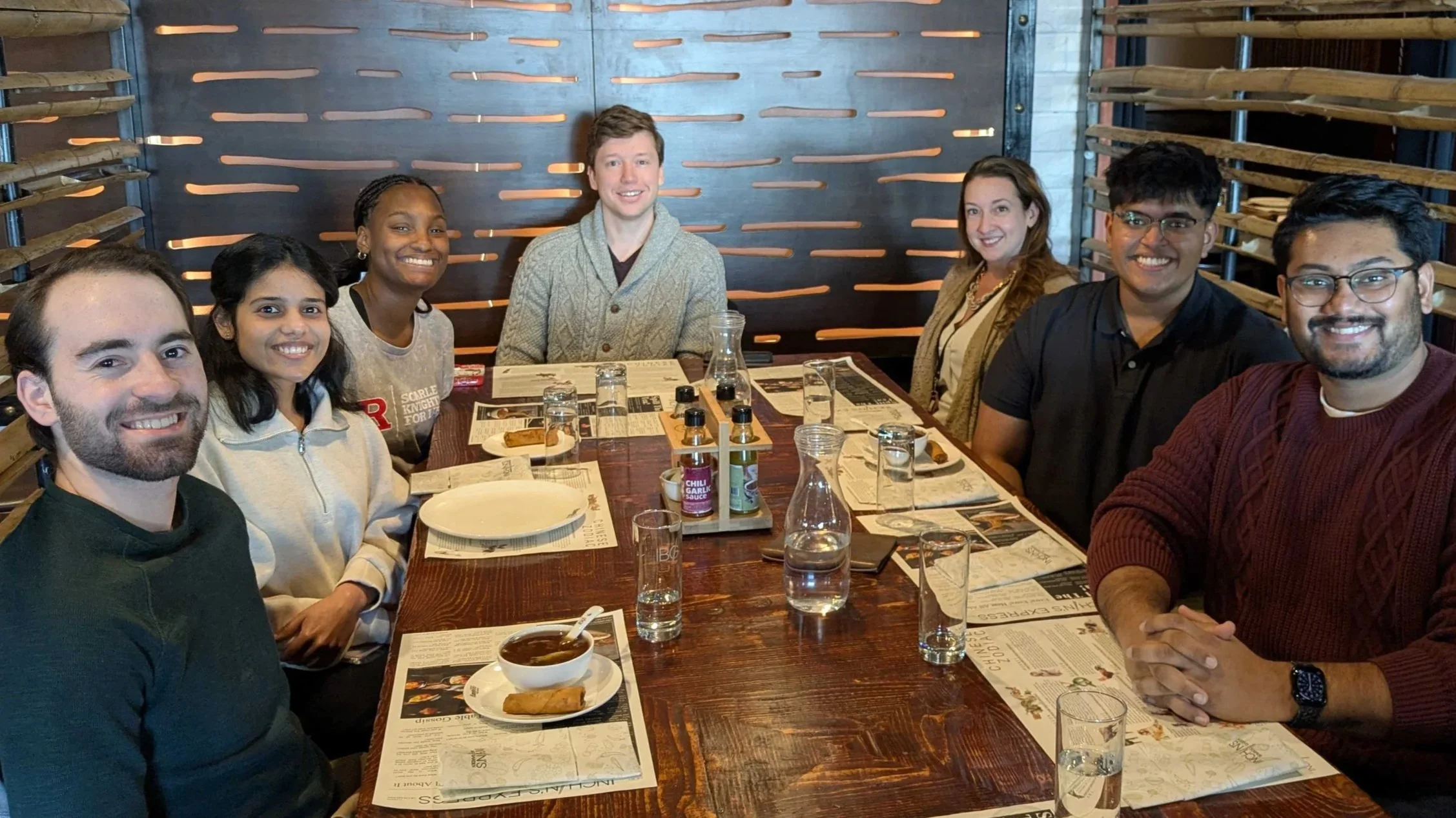 Group of seven diverse people seated around a wooden table in a restaurant, smiling at the camera, with menus and drinks on the table.