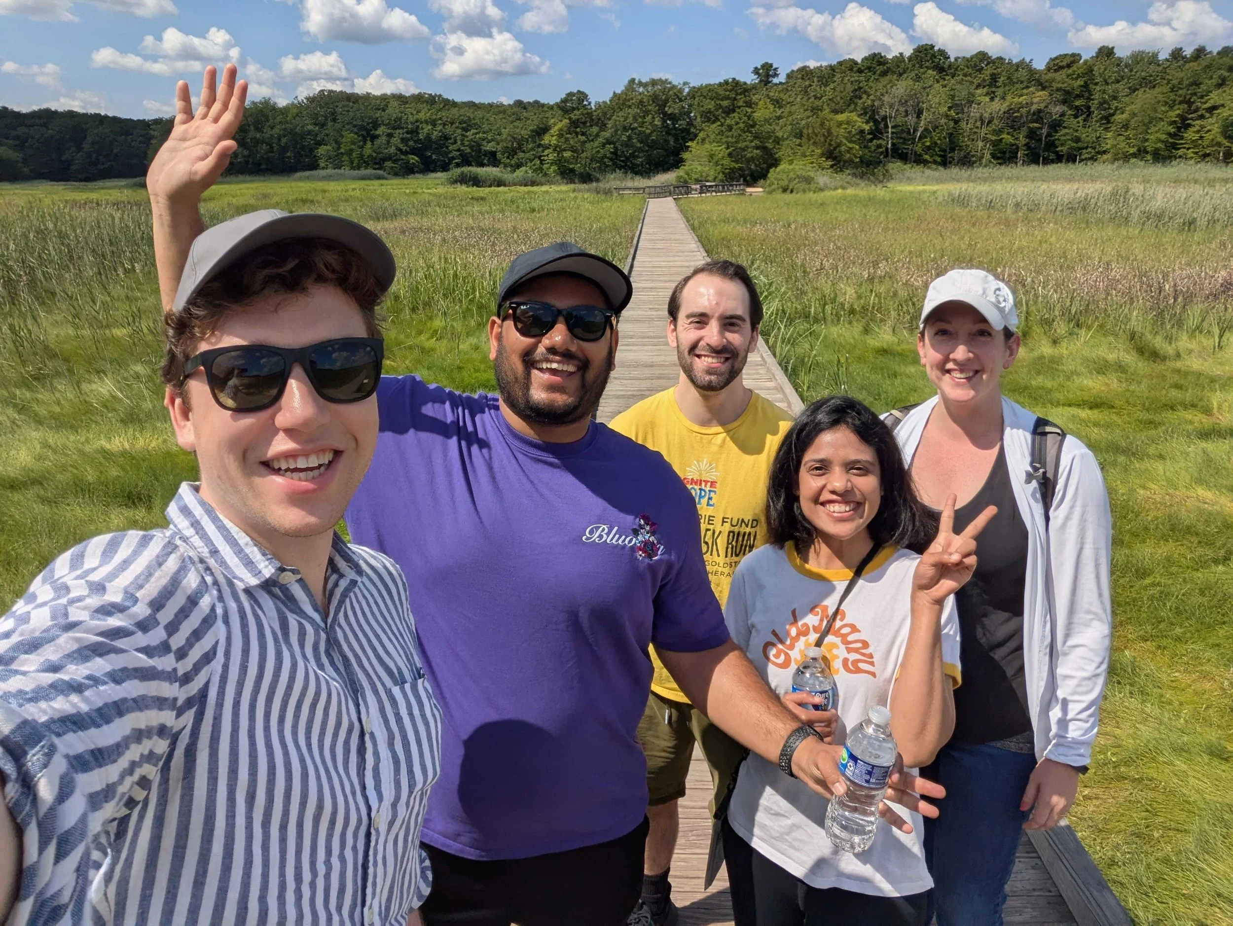 Group of five smiling people taking a selfie on a wooden bridge in a grassy park with trees and a cloudy sky in the background.