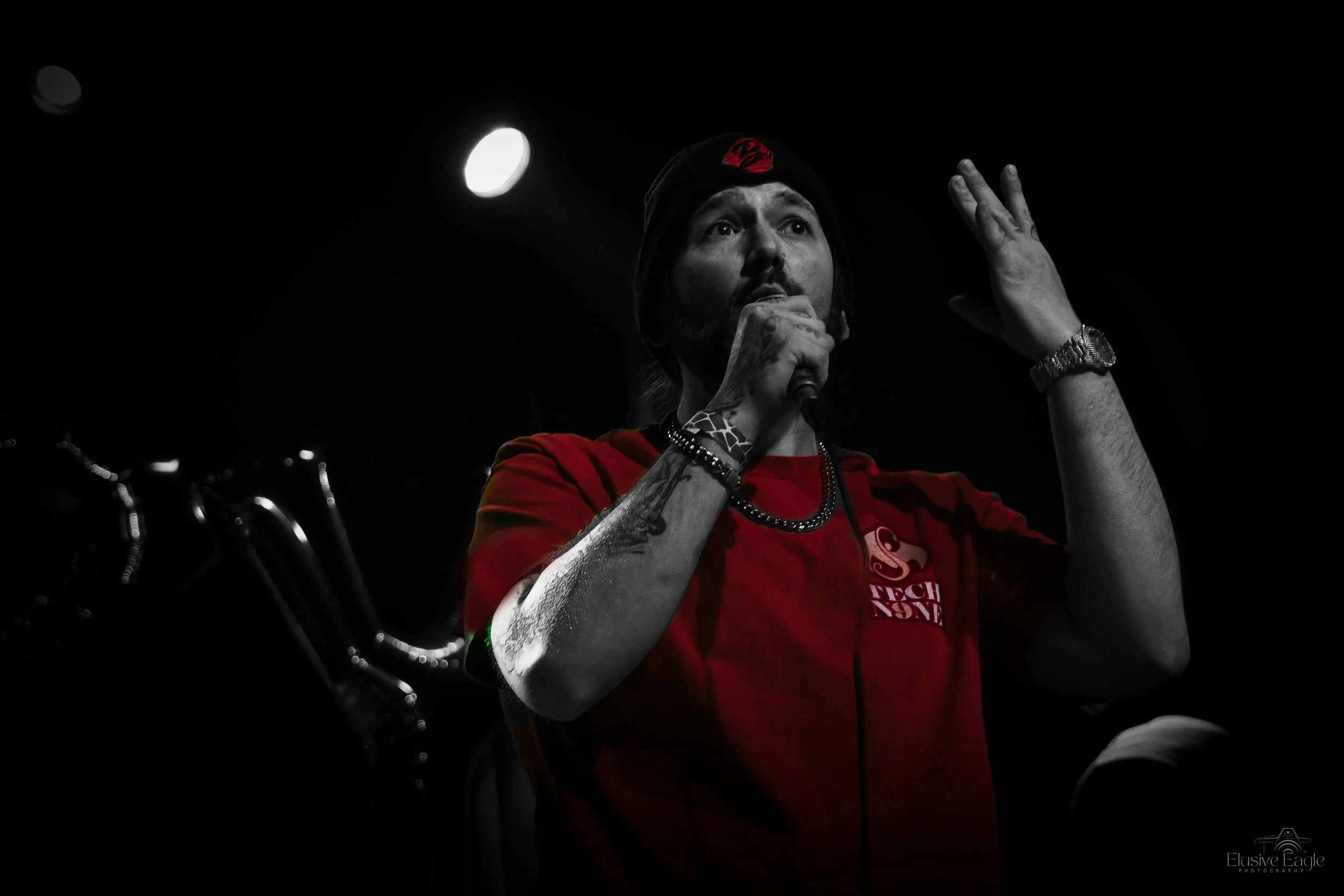 Performer on stage with a microphone, wearing a red shirt and a beanie, with expressive hand gestures under spotlight.