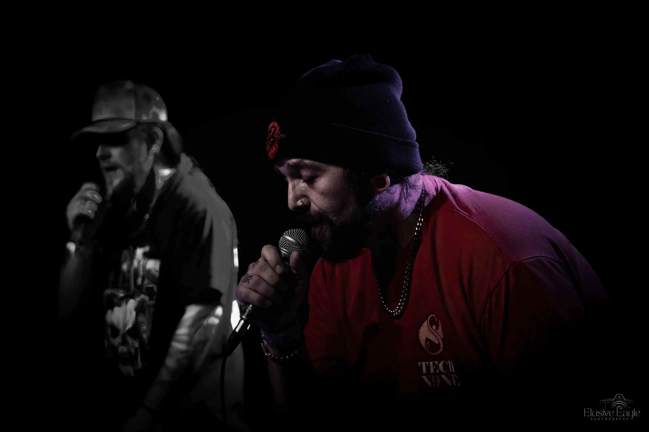 Two musicians performing on stage, one in a red shirt with "Tech N9ne" logo, wearing a beanie and holding a microphone. The background is dark, focusing the light on their silhouettes.