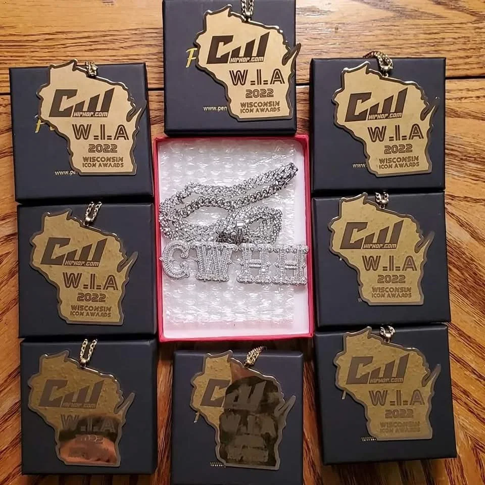 Six black boxes with gold 'WI' awards and a necklace with a silver 'CHY' pendant and another chain lying on a table. One box is open, revealing a silver chain necklace in a red box.