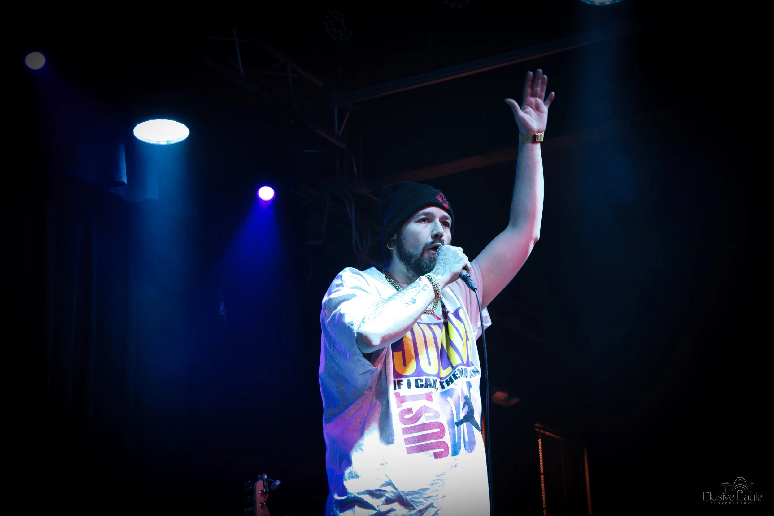 Rapper performing on stage with microphone, wearing a beanie and graphic t-shirt, under stage lights.