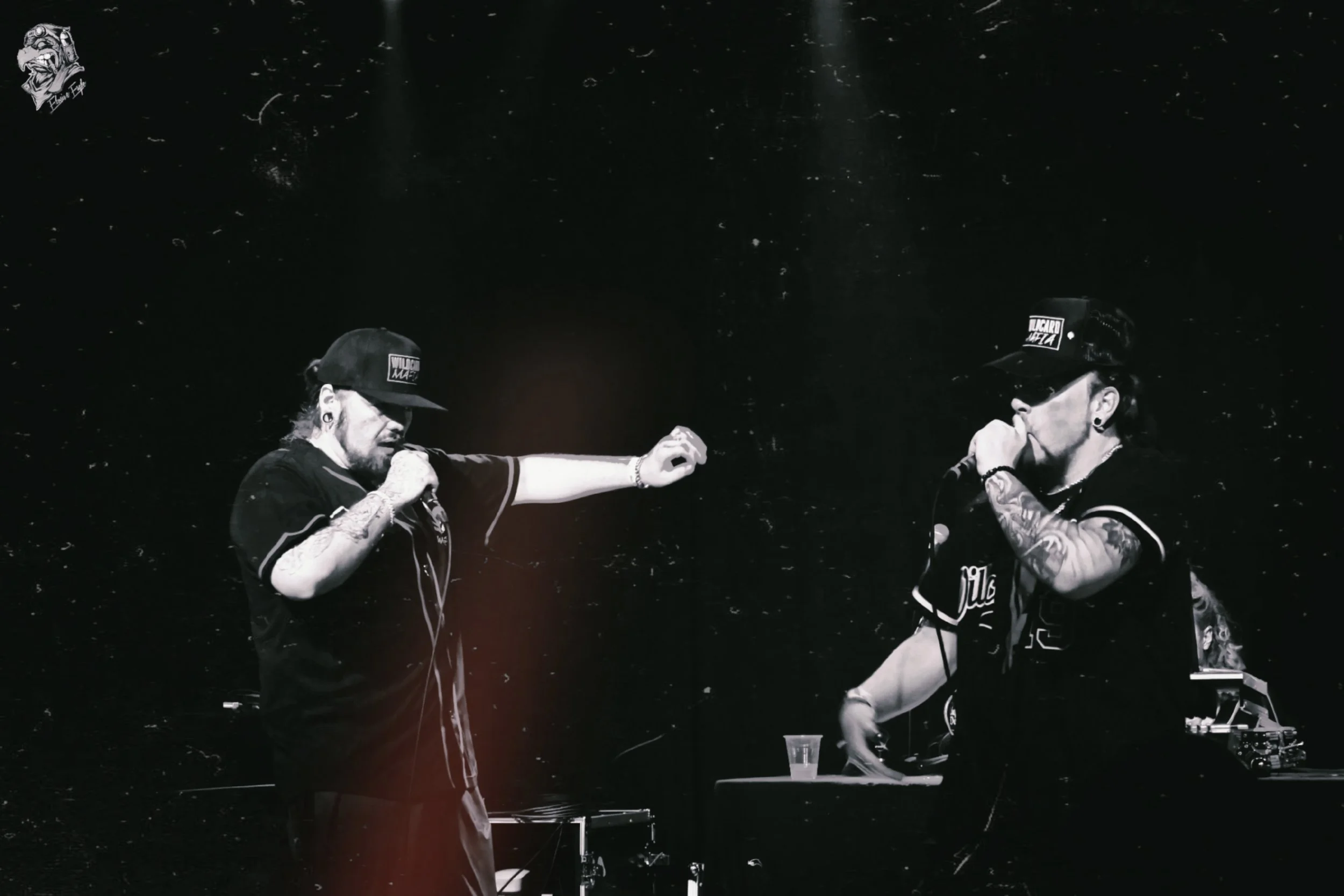 Two tattooed performers with microphones on stage, wearing baseball caps, in a monochrome photo