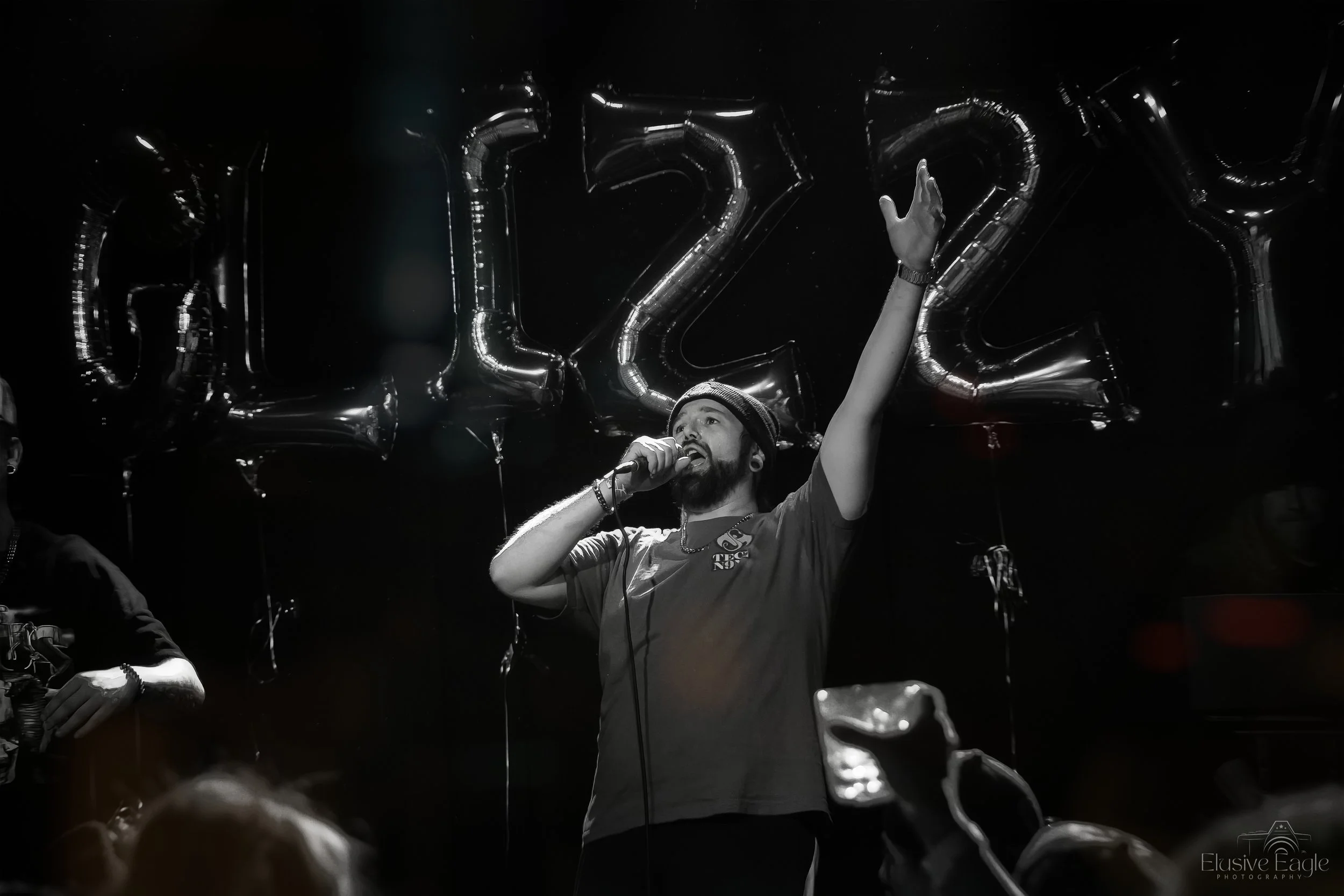 A person singing on stage with a microphone, surrounded by balloons spelling "GLIZZY" in the background.