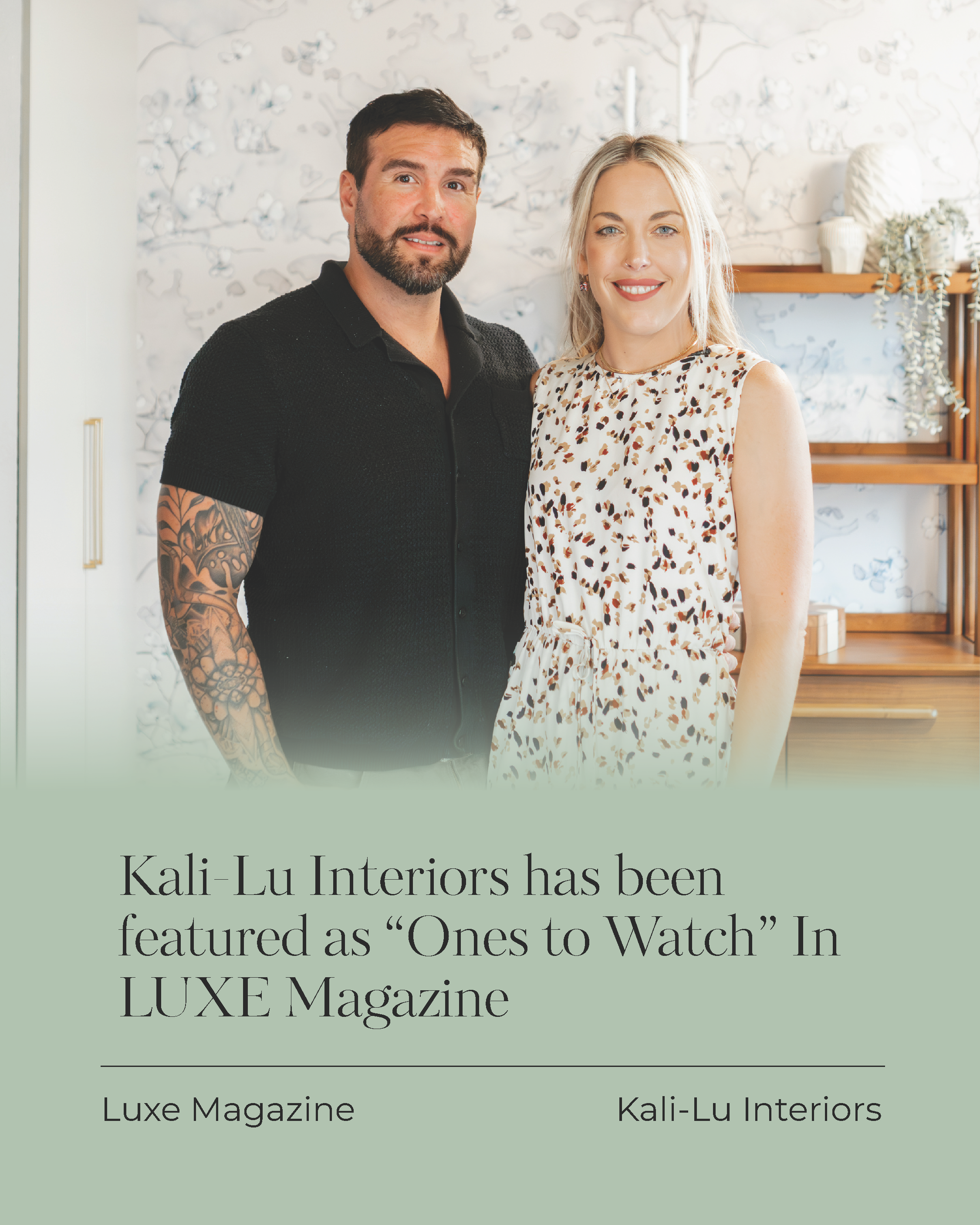 A man with a beard and tattooed arm and a woman with blonde hair smiling, standing indoors in front of a decorative wall with floral wallpaper and a wooden shelf with white vases and plants.