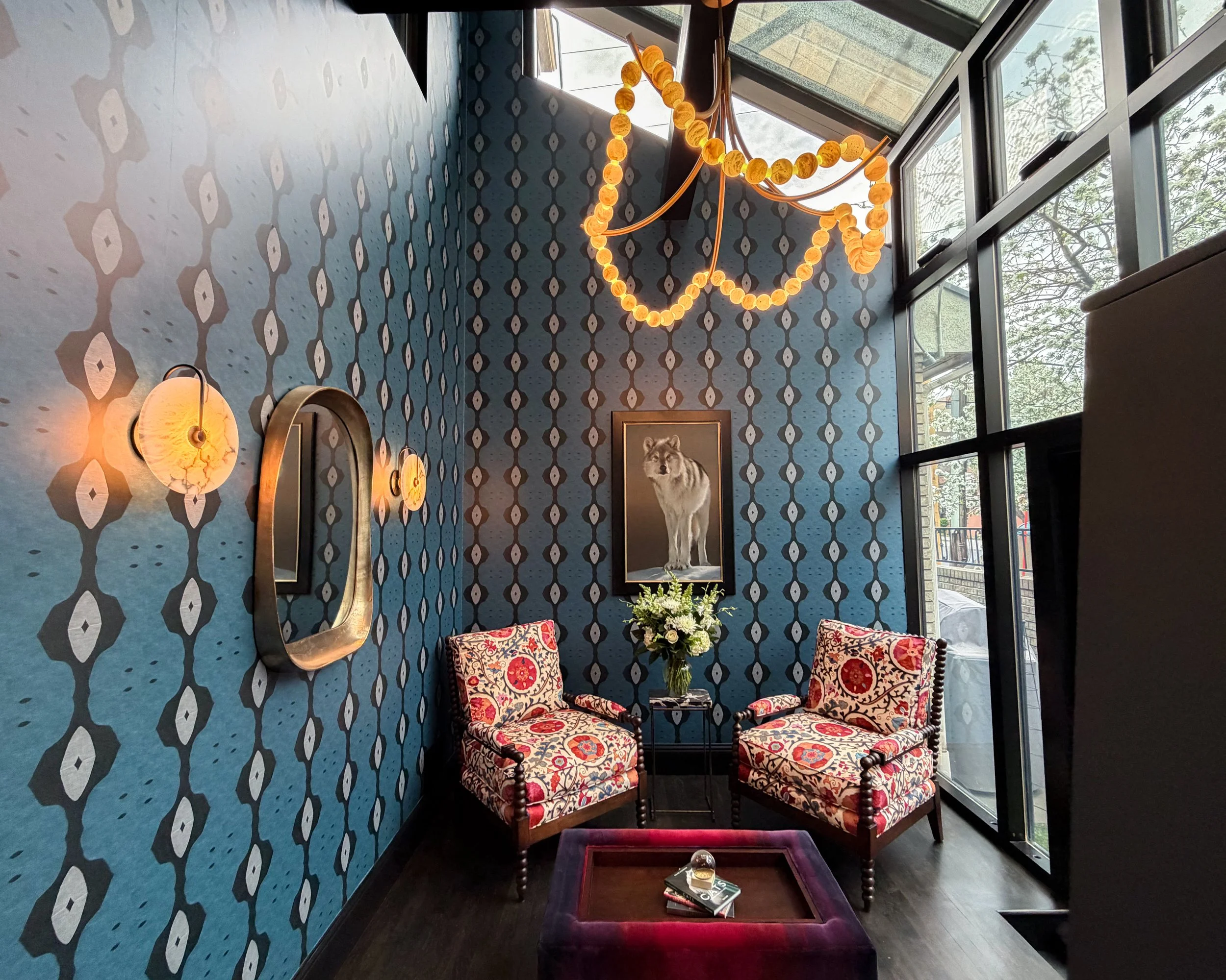 A sunroom adorned with printed blue, black, and ivory wallpaper with two pink and orange printed chairs centered around a wolf photo and purple and red ottoman. The room is lit with the natural light from the windows and a golden glow from fixtures.