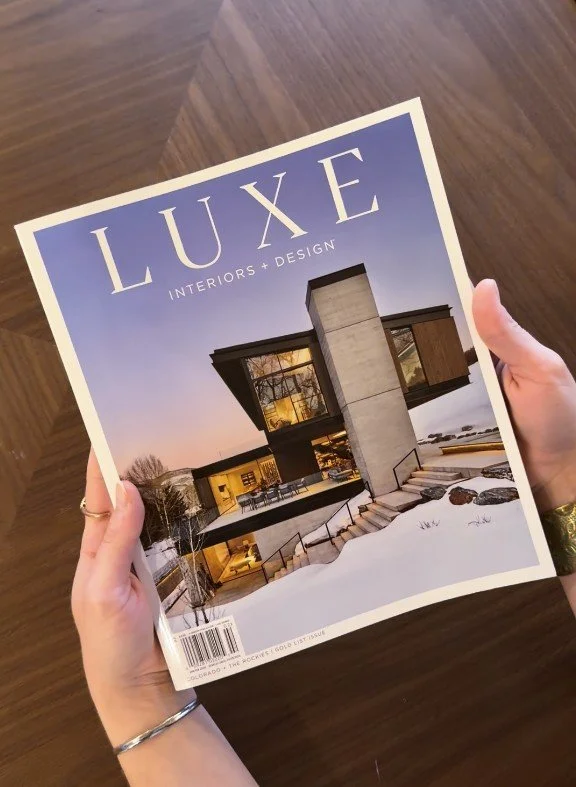 A person holding a magazine titled 'LUXE Interiors + Design' showing a modern house exterior with snow-covered ground.