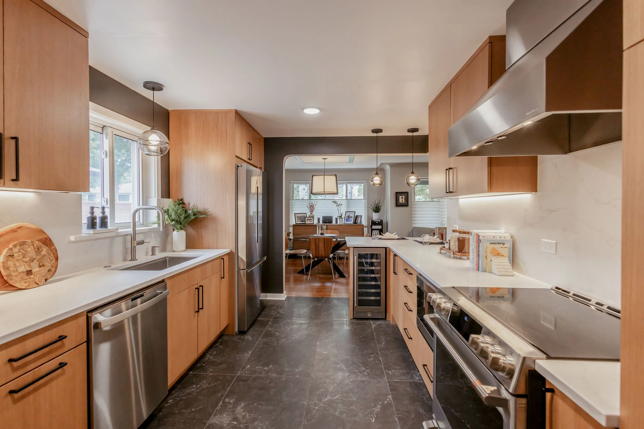 Modern kitchen with wooden cabinets, black marble flooring, stainless steel appliances, and a view into the dining area with a wooden table and chairs, pendant lights, and large windows.