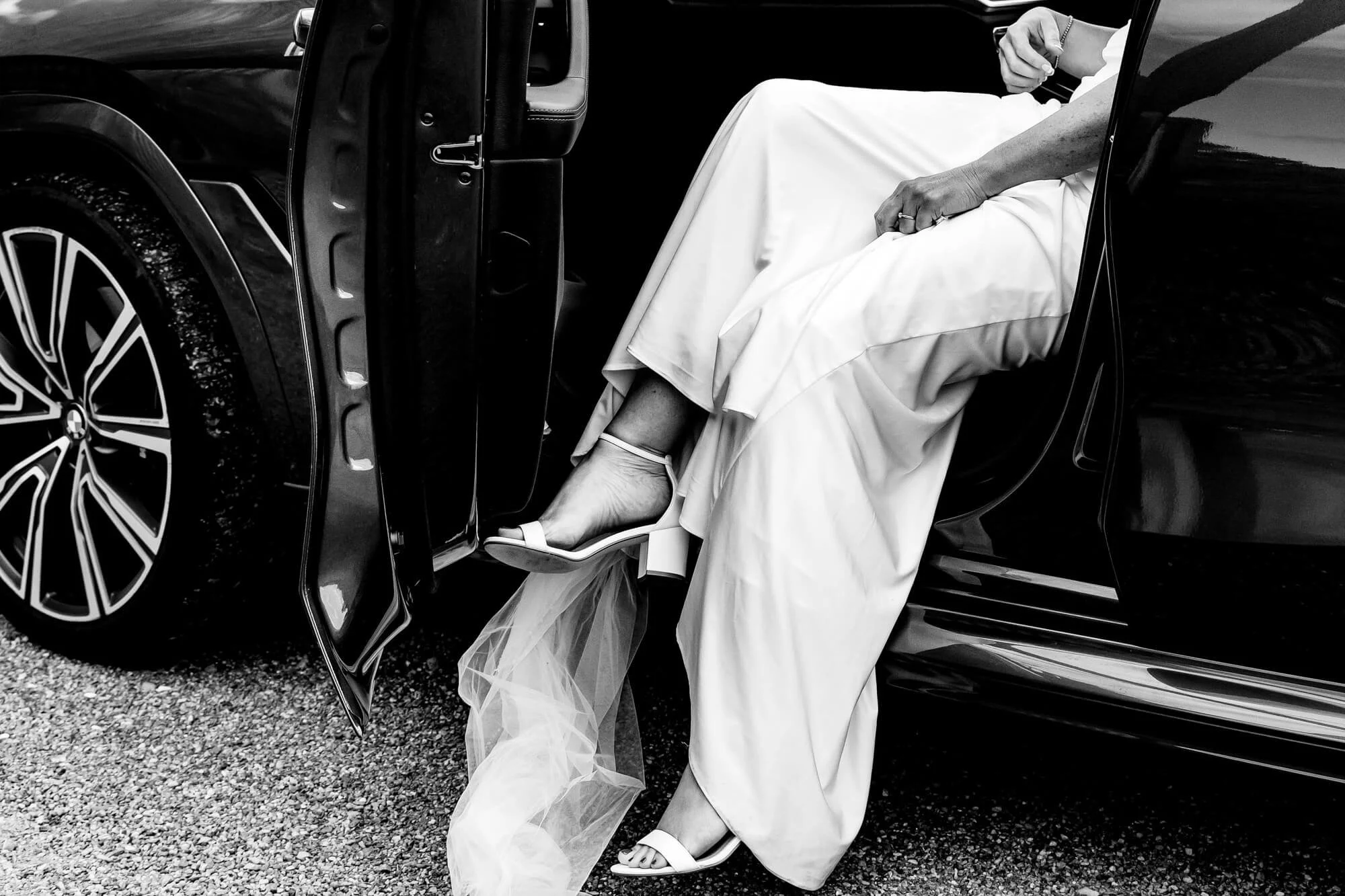 A bride in white wedding dress and heels stepping out of a black luxury car.