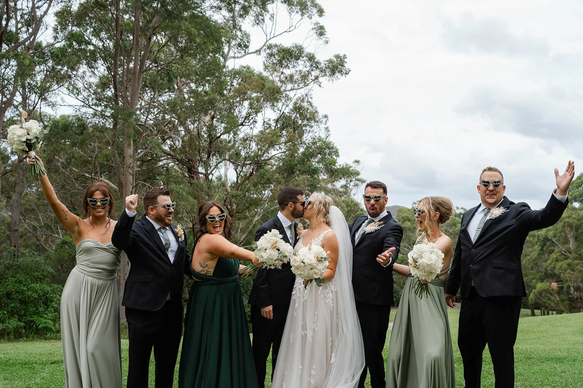 Group of wedding guests, including bride and groom, celebrating outdoors on a cloudy day, wearing sunglasses, with some holding bouquets and others raising their hands in celebration.