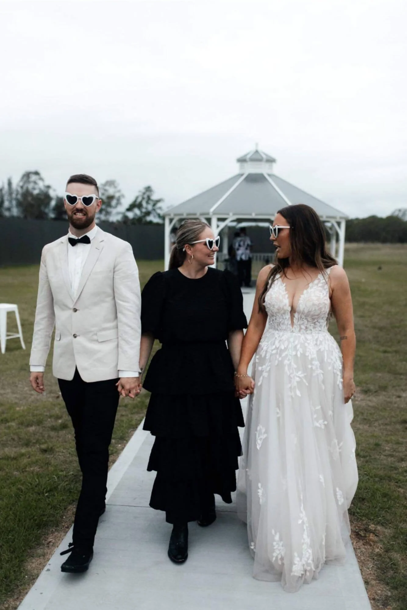 Three people holding hands, two women and one man, walking outdoors during a wedding celebration. The woman on the right is in a white wedding dress, the man on the left is in a white jacket with black pants and bow tie, and the woman in the middle i
