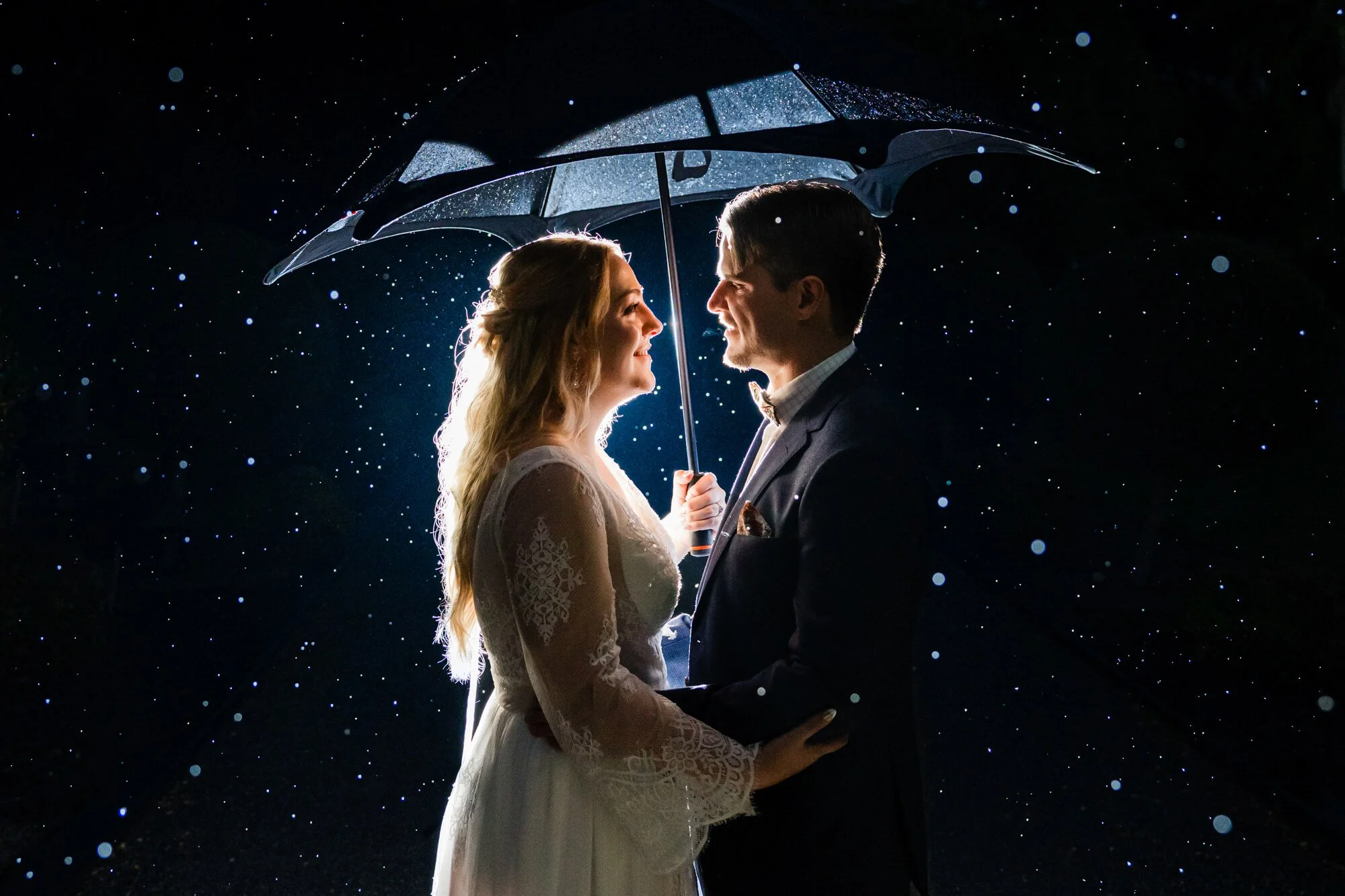 A couple in wedding attire standing close together under an umbrella at night, with rain falling around them. They are looking at each other and smiling.