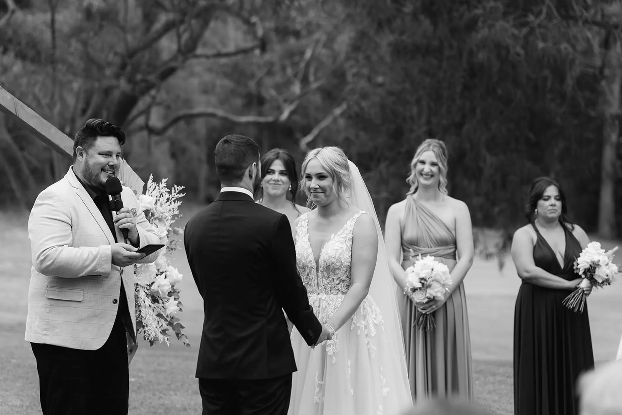 A black and white photograph of a wedding ceremony outdoors, with the bride and groom holding hands, facing each other, with the officiant speaking into a microphone. Three women in dresses are standing behind them, smiling and holding bouquets.