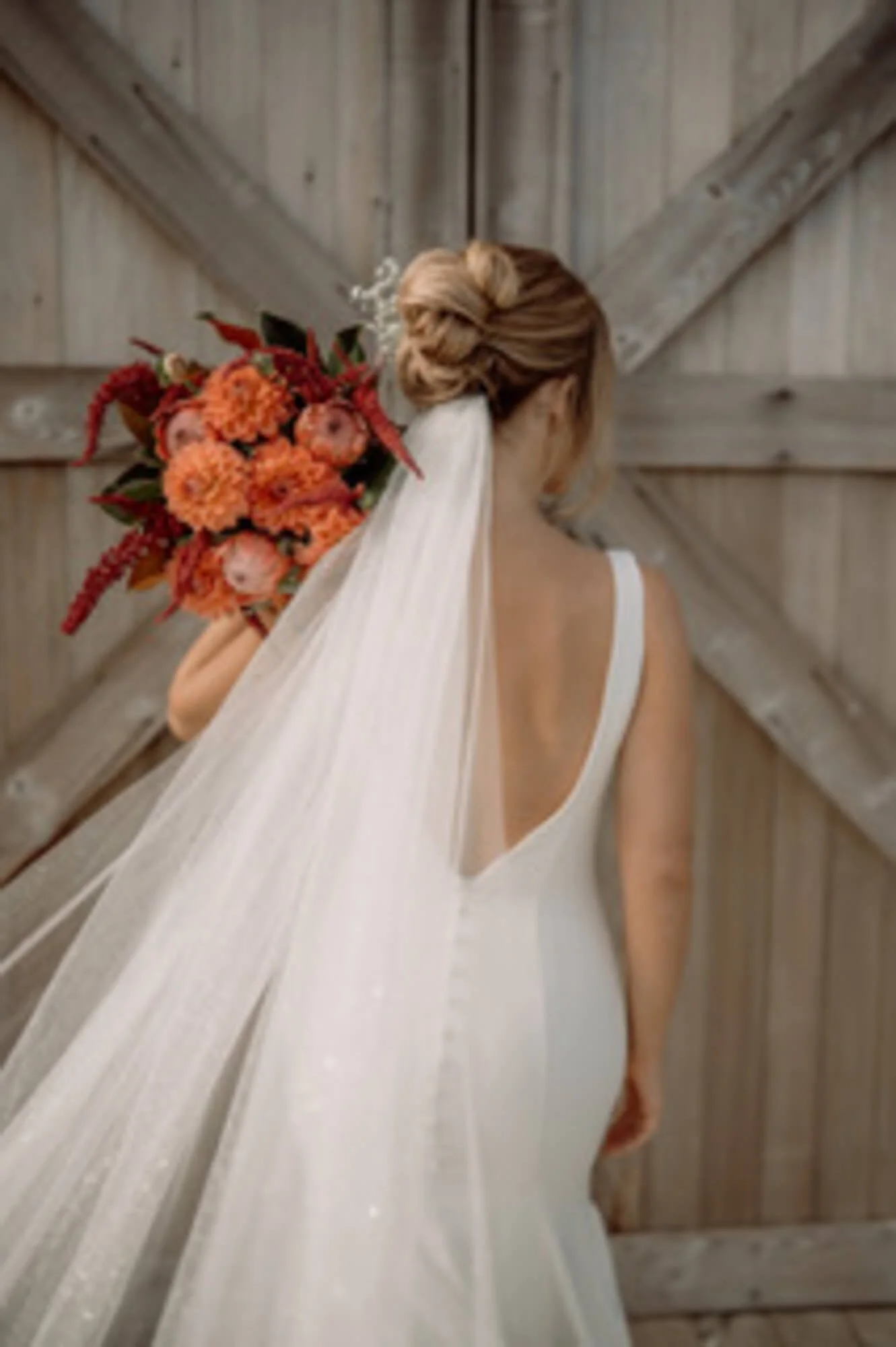 Bridal portrait of a woman in a white wedding gown with a low back, holding a bouquet of orange, red, and pink flowers, with her hair styled in an updo and a veil hanging down her back, against a rustic wooden barn door.