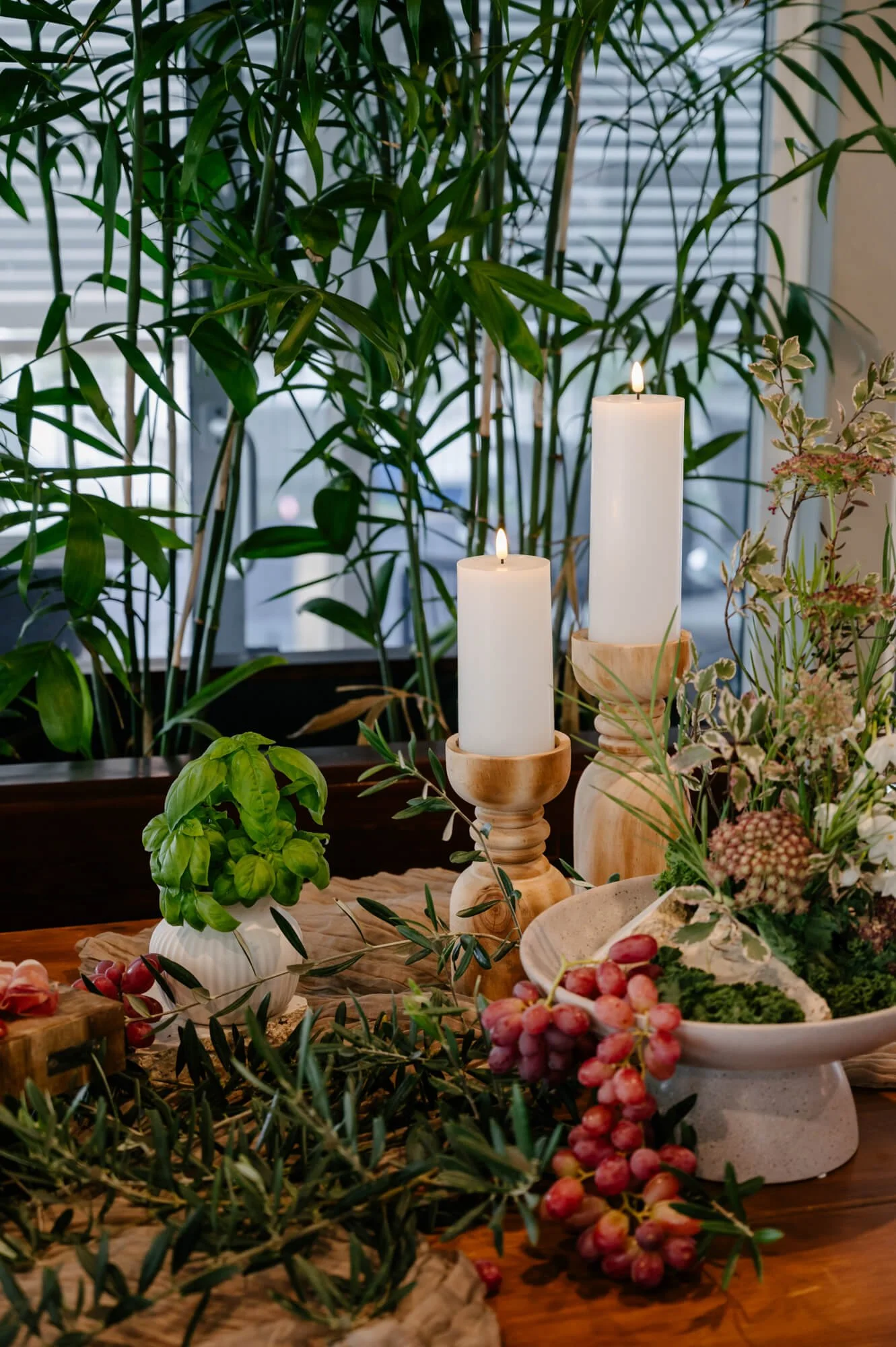 A table decorated with white candles on wooden holders, a potted basil plant, pink grapes, and flowers, with large green houseplants and blinds in the background.