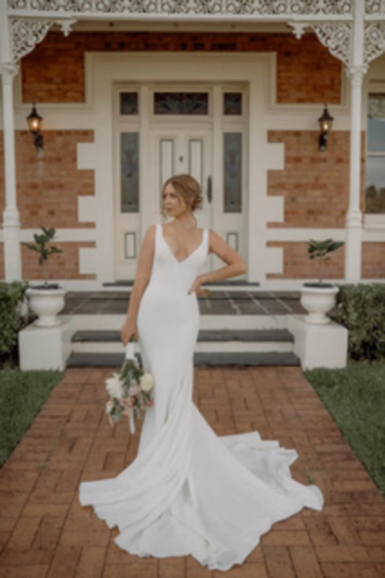 A bride in a white wedding dress holding a bouquet stands in front of a brick house with white decorative trim and potted plants on the steps.
