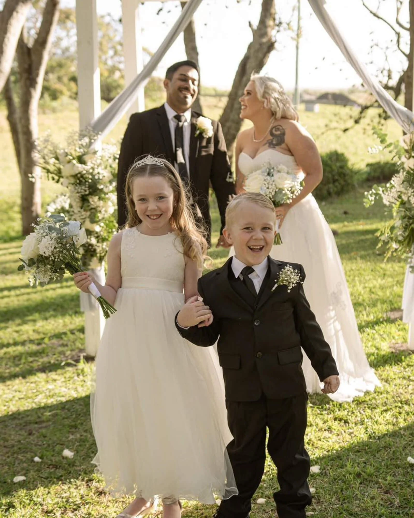 A wedding ceremony outdoors with a bride and groom, and two children in formal attire, smiling and holding flowers.