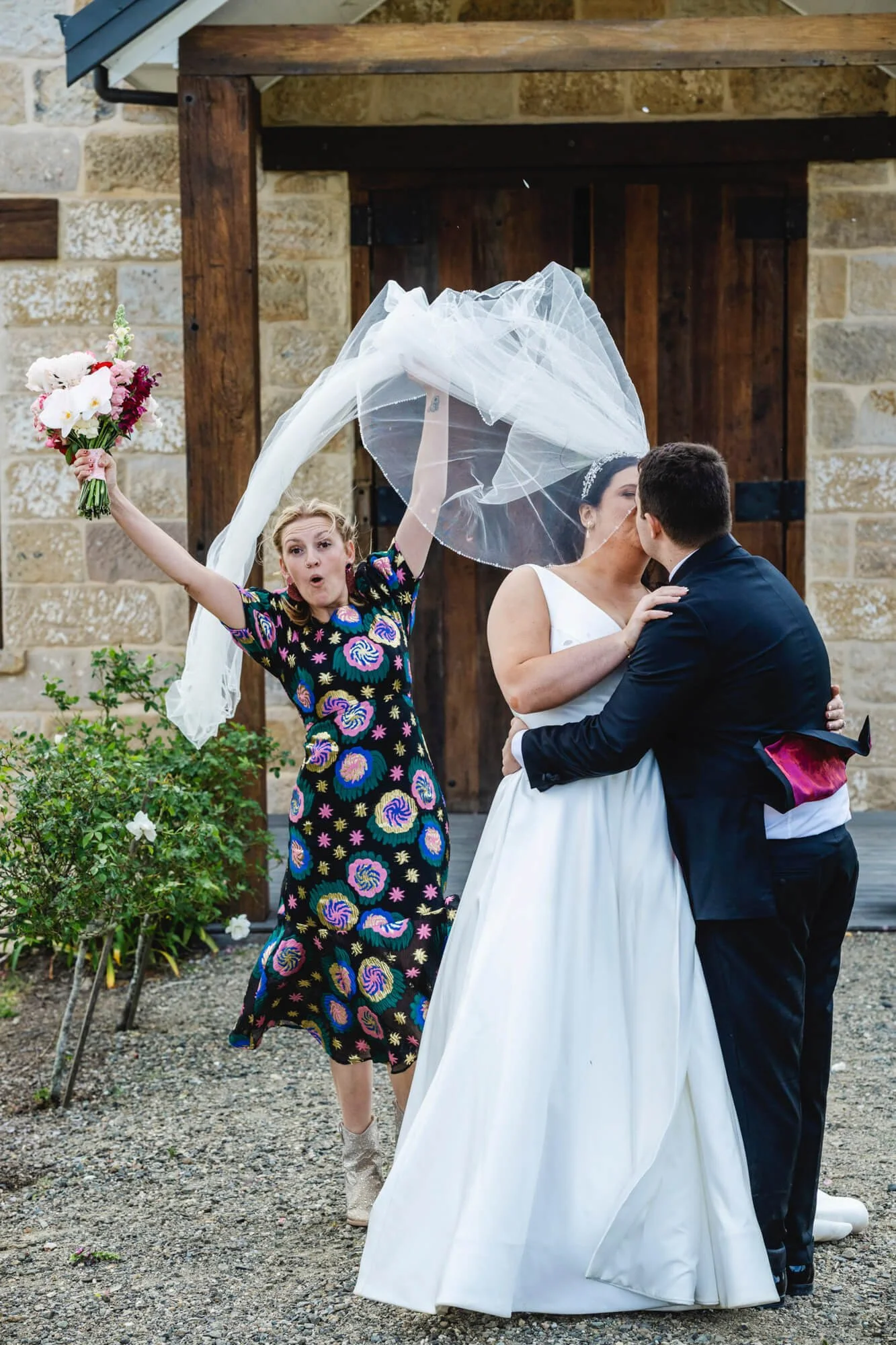 A bride and groom share a kiss outdoors, while a woman in a colorful dress excitedly throws her veil into the air, holding a bouquet of flowers.