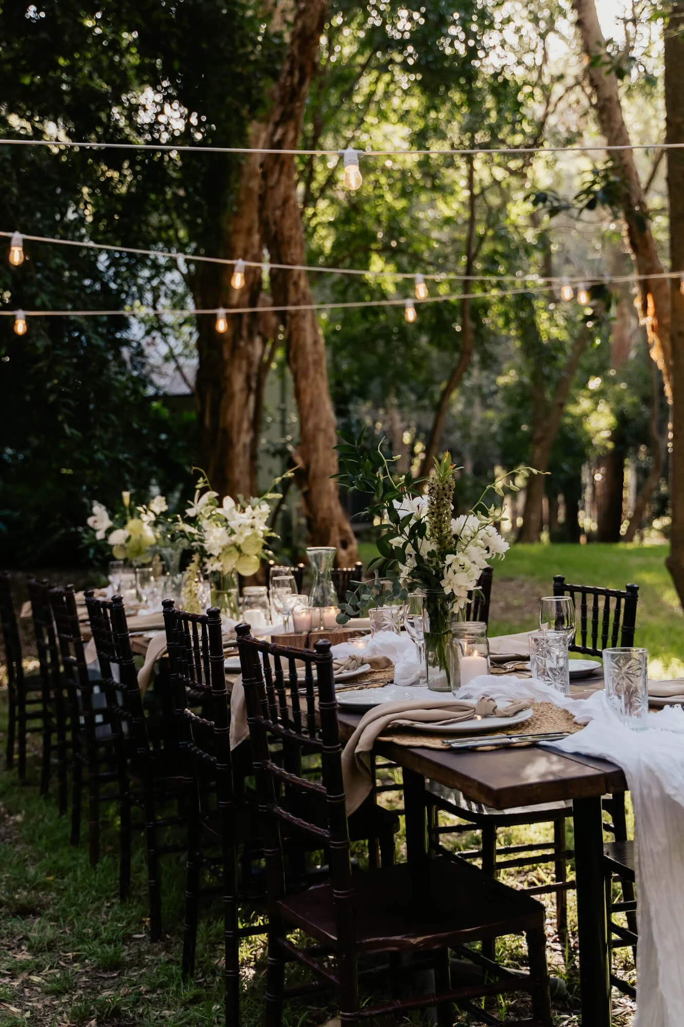 An outdoor dining table set for a gathering, decorated with floral centerpieces, candles, and tableware, under string lights in a wooded area.