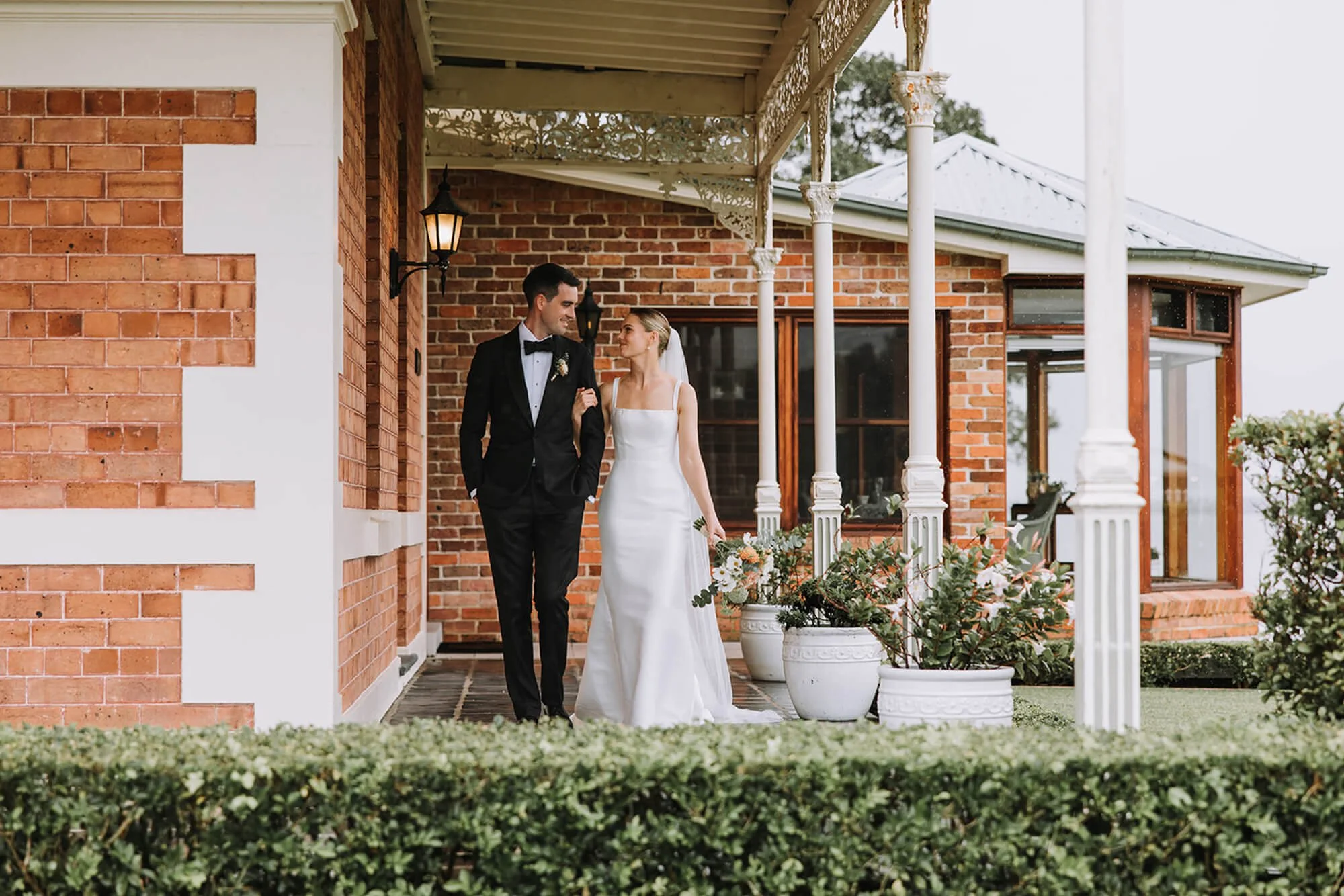 A bride and groom walking arm in arm on a porch of a brick house. The groom is wearing a black tuxedo and the bride is in a white wedding gown with flowers in her hair. The porch has white columns and potted plants, with a background of trees and a c