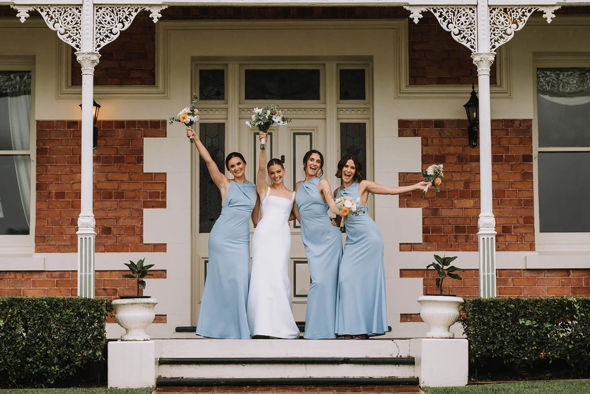 Four women standing on a porch, celebrating a wedding, with three holding bouquets. The woman in the center wears a white dress, while the other three women wear matching blue dresses. They are smiling and celebrating with arms raised.