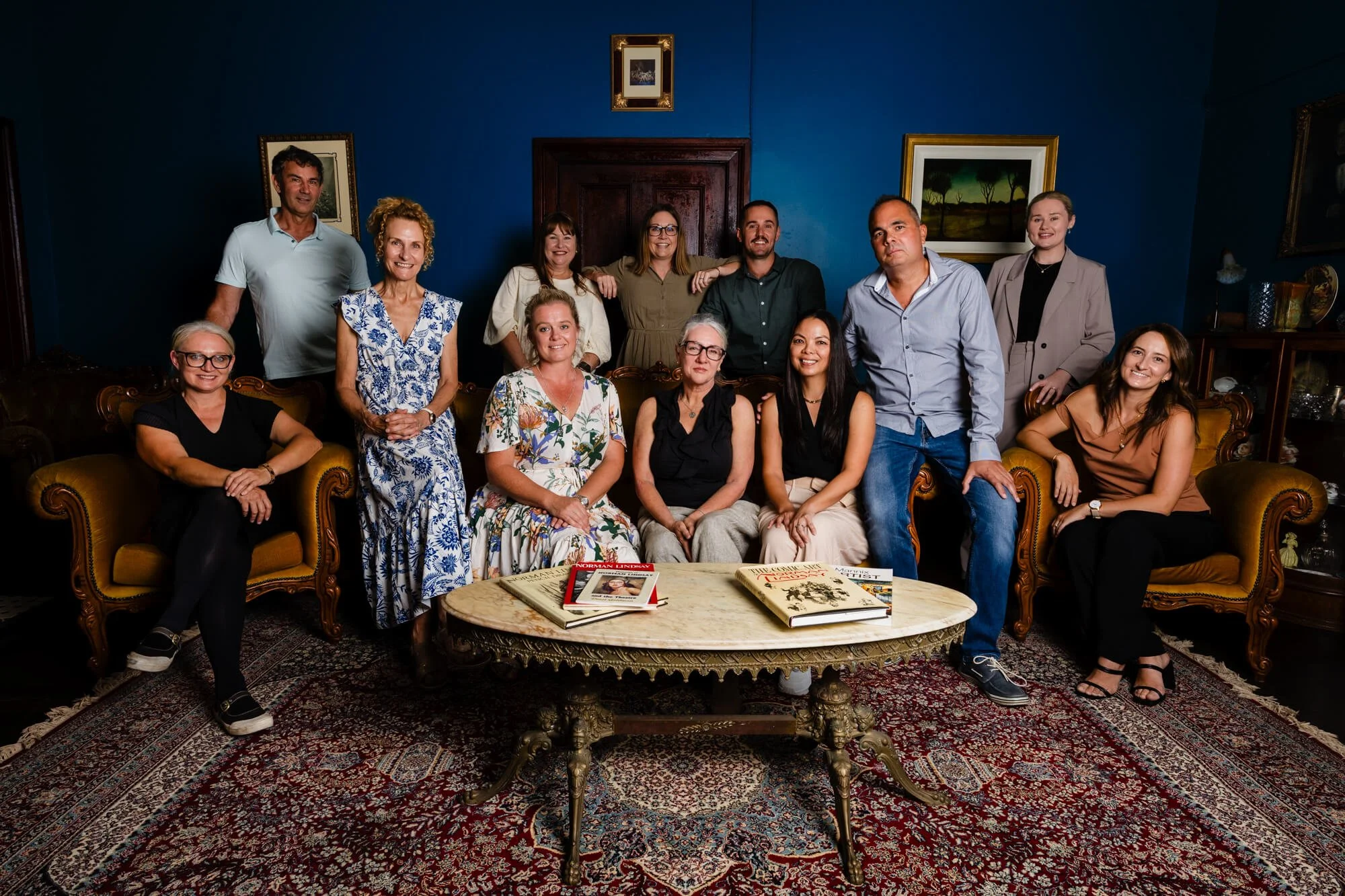A group of fifteen people posed together in a living room, sitting and standing around a vintage-style coffee table, with dark blue walls and framed paintings in the background.