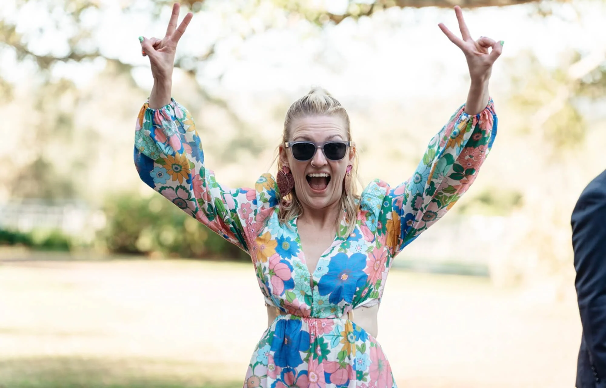 A woman wearing sunglasses and a colorful floral dress, smiling and making peace signs with both hands outdoors.