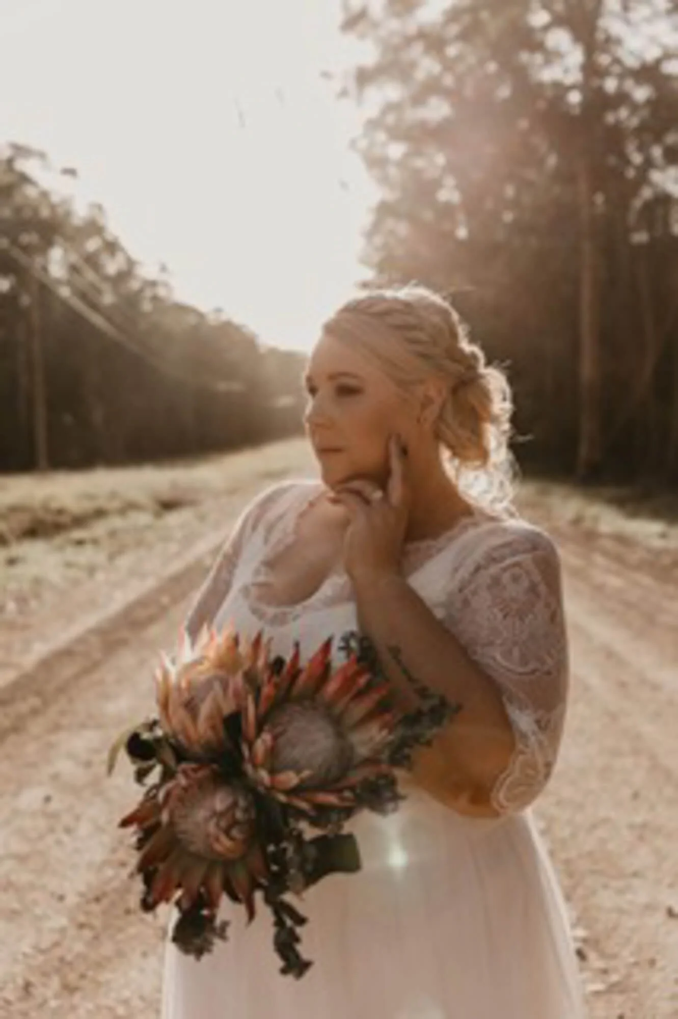 A woman in a wedding dress holding a bouquet of flowers on a dirt road with trees in the background, sunlight shining brightly.