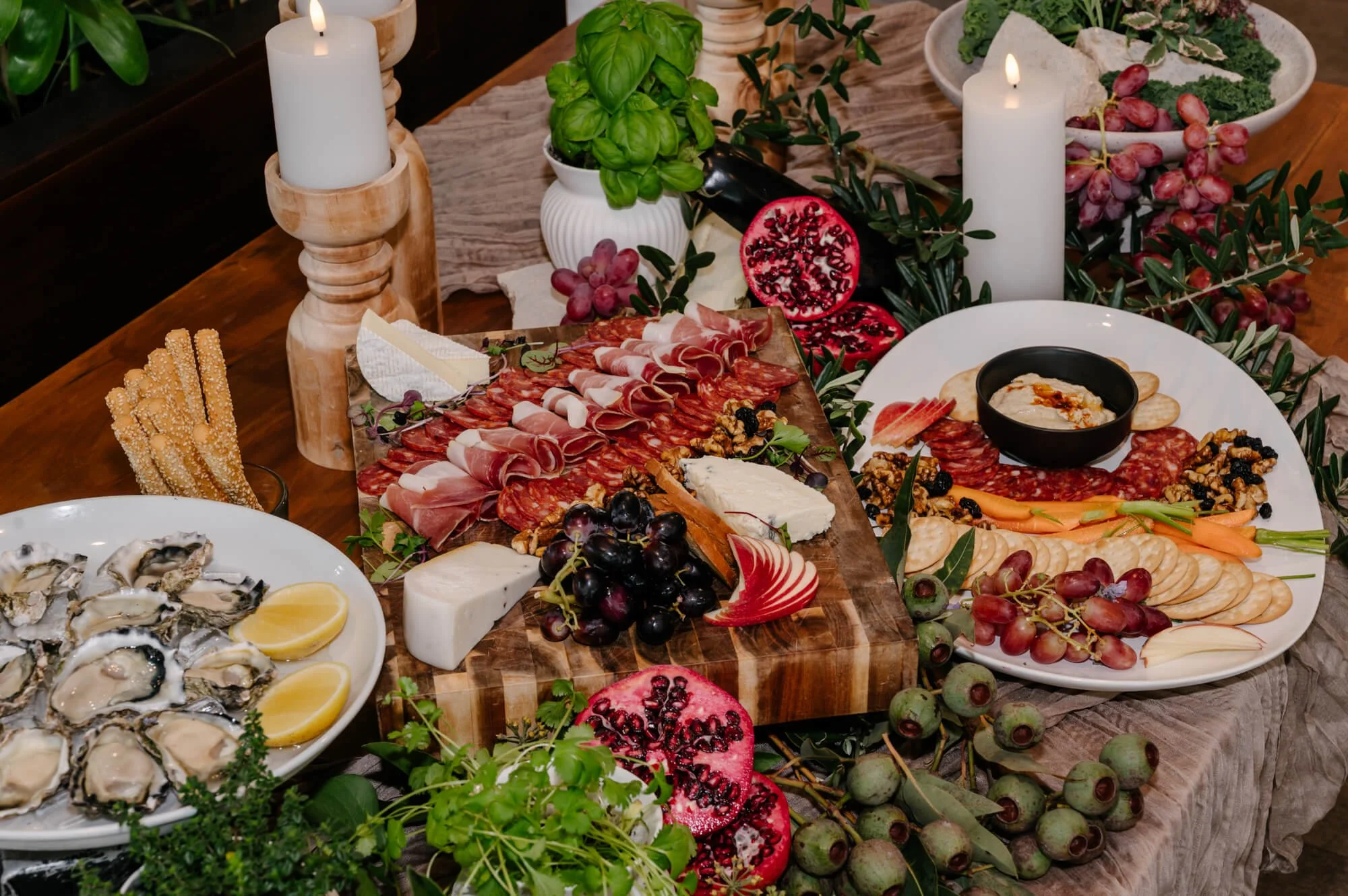 Charcuterie board with meats, cheeses, grapes, and vegetables, surrounded by candles, pomegranate, oysters, breadsticks, and greenery on a wooden table.