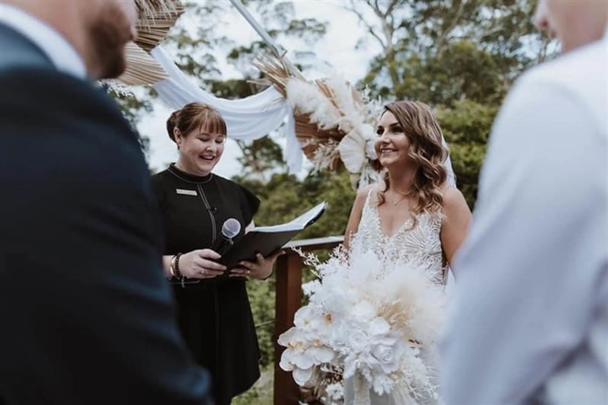 A bride smiling during her outdoor wedding ceremony, standing in front of an officiant woman reading from a book, with floral decorations and trees in the background.