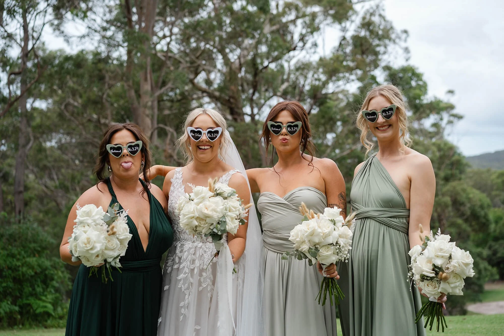 Four women standing outdoors on a cloudy day, wearing bridesmaid dresses and holding bouquets of white flowers. They are making funny faces and wearing black sunglasses shaped like lips with white writing on the lenses that say 'Bride' or 'Groom' and