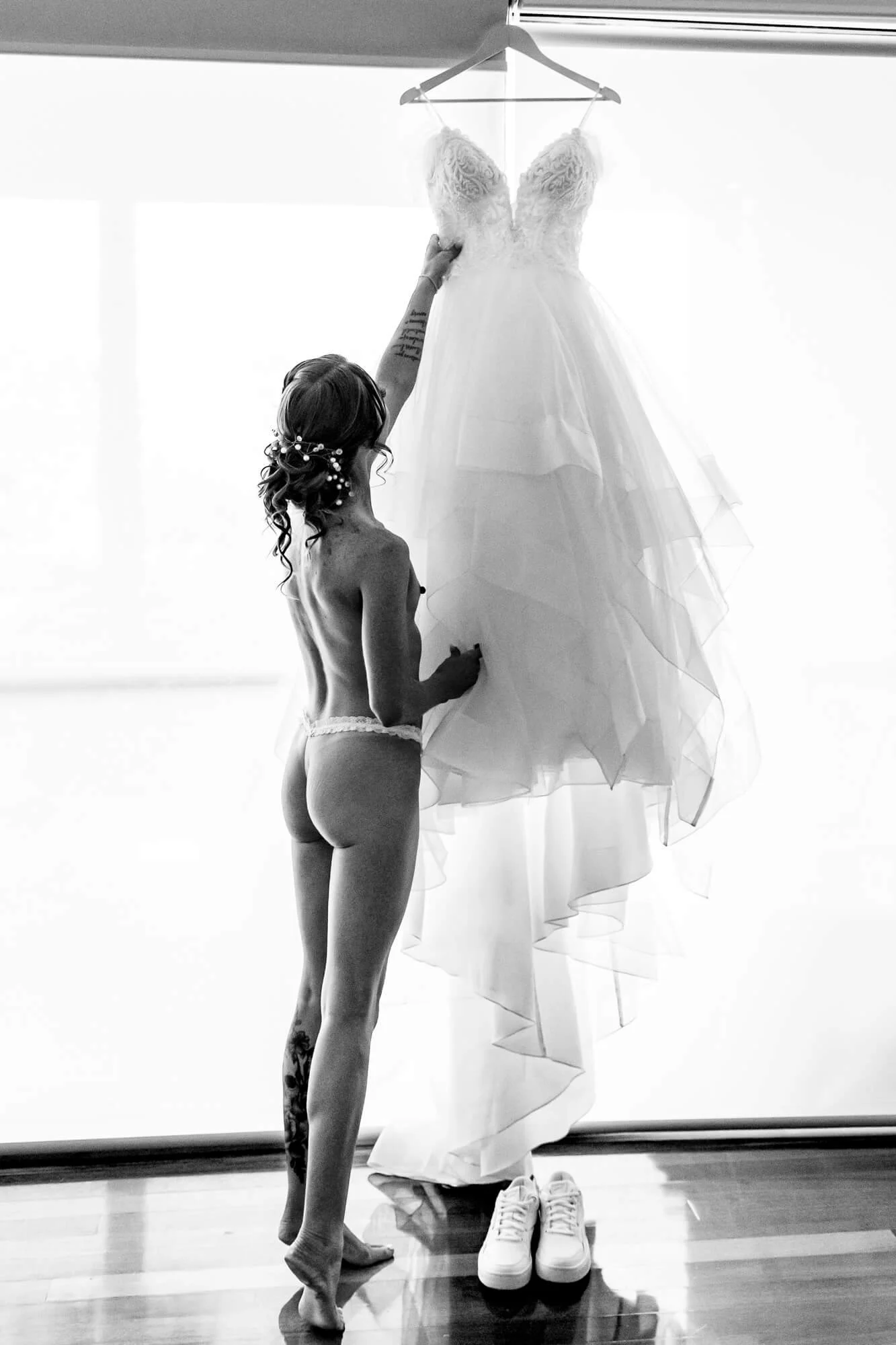 Black and white photo of a woman with tattoos and long hair, wearing only underwear, reaching up to touch a wedding dress hanging from a hanger. The wedding dress is off the ground, with shoes placed underneath it, in a bright room with hardwood floo