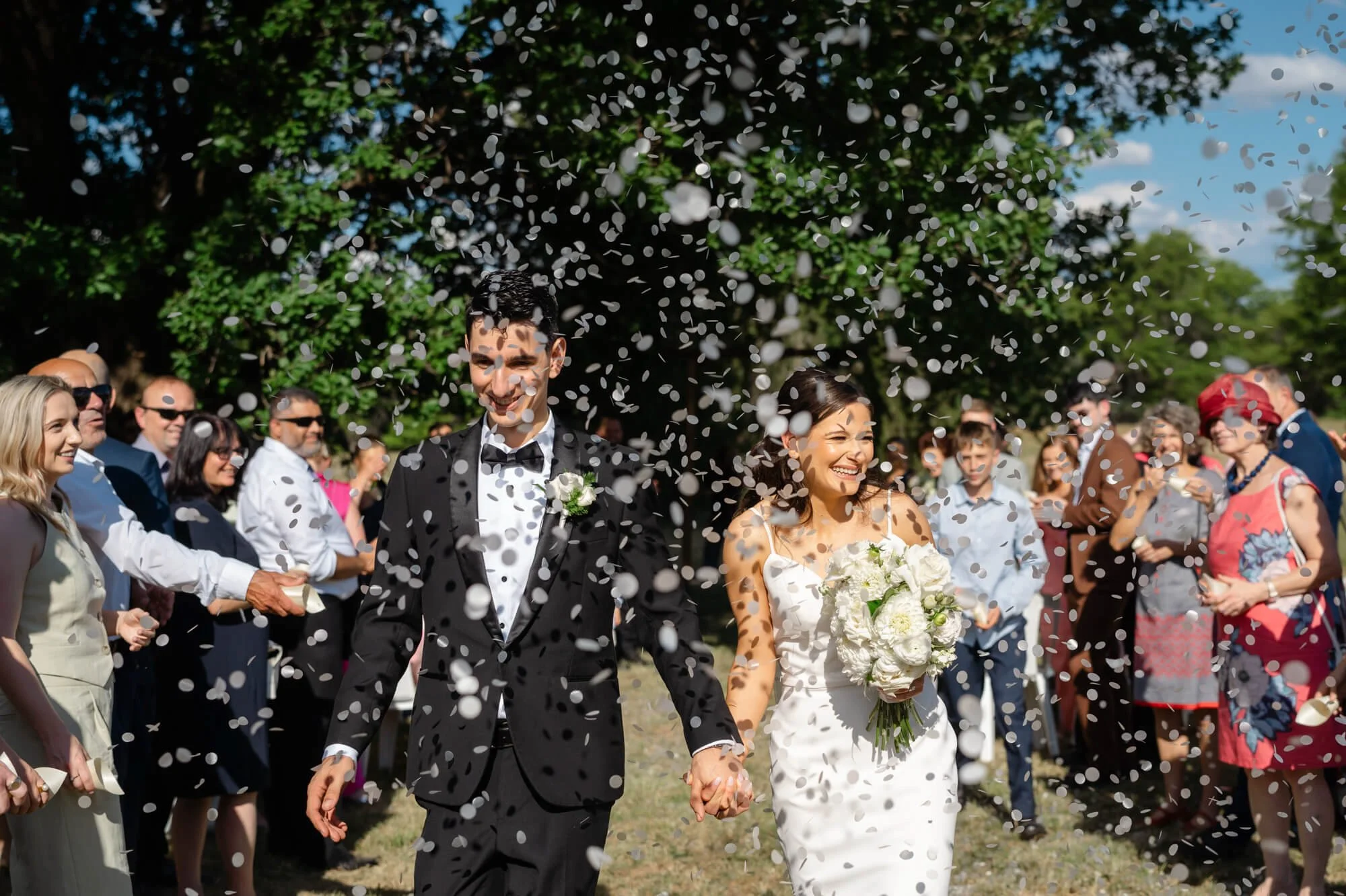 A bride and groom holding hands walking through a shower of confetti with friends and family in the background outdoors.