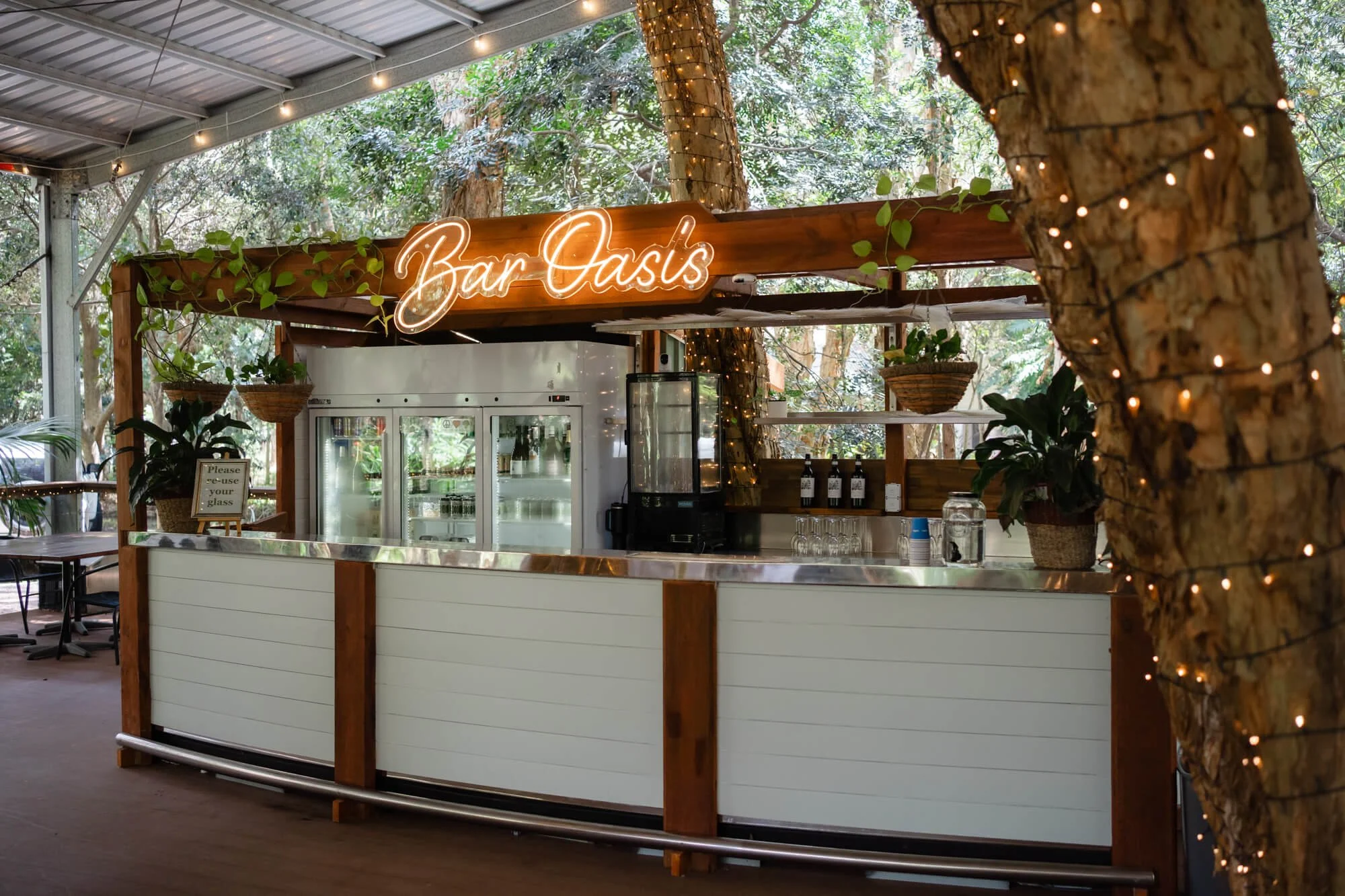 An outdoor bar area called 'Bar Oasis' with a neon sign, surrounded by trees with string lights, and a white counter with plants and glassware.